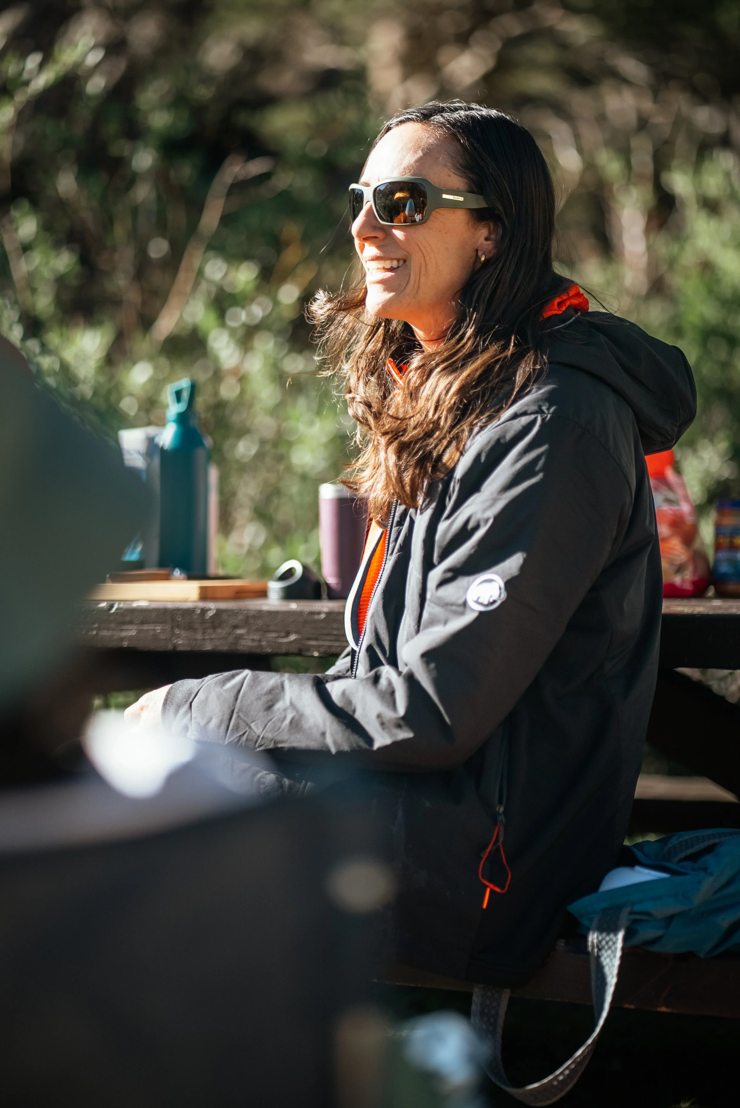 Woman with long wavy hair wearing sunglasses and a black jacket, sitting outdoors at a picnic table, smiling in sunlight.