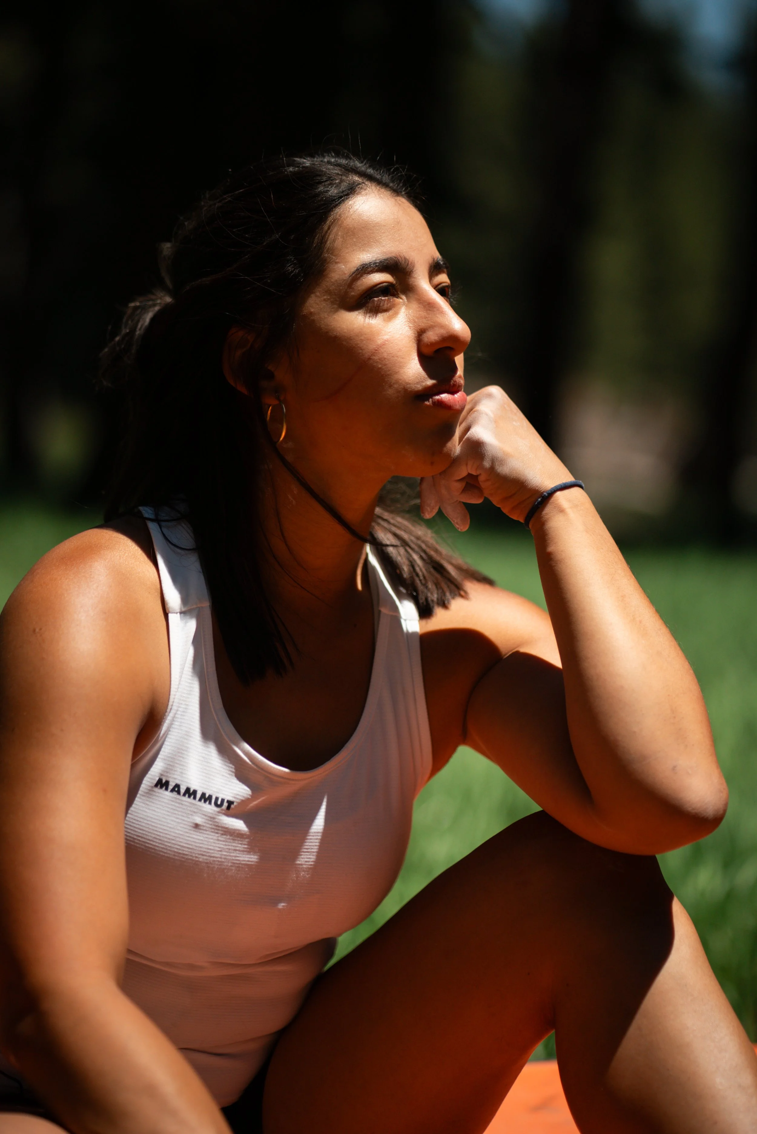 A woman sitting outdoors on grass with trees in the background, wearing a white athletic top with a 
