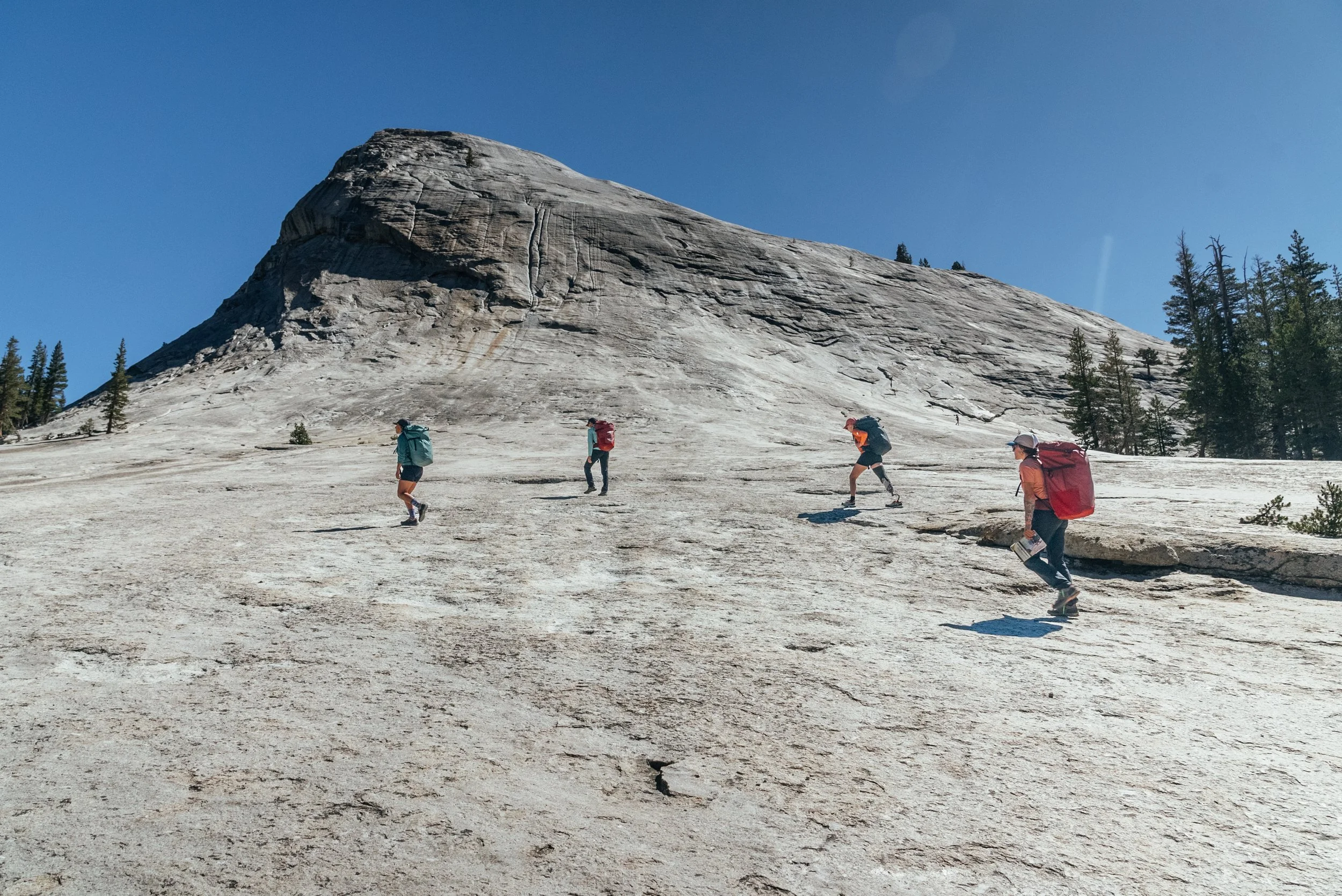 Four hikers with backpacks ascending a rocky, sloped mountain under a clear blue sky, with sparse trees on the slope.
