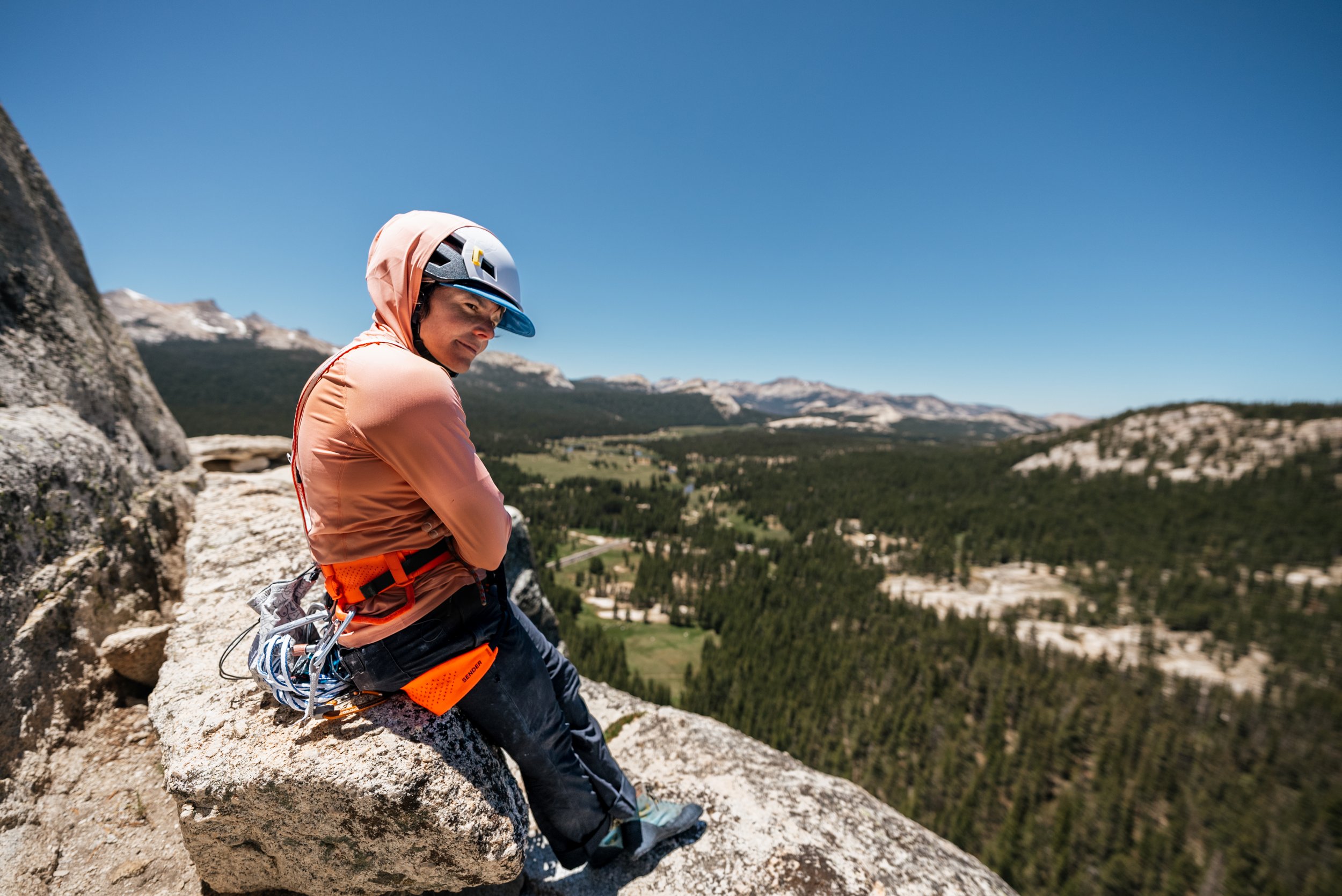 A rock climber sitting on a large rock ledge, wearing a helmet, orange jacket, and climbing gear, overlooking a vast landscape of forested hills and mountains under a clear blue sky.