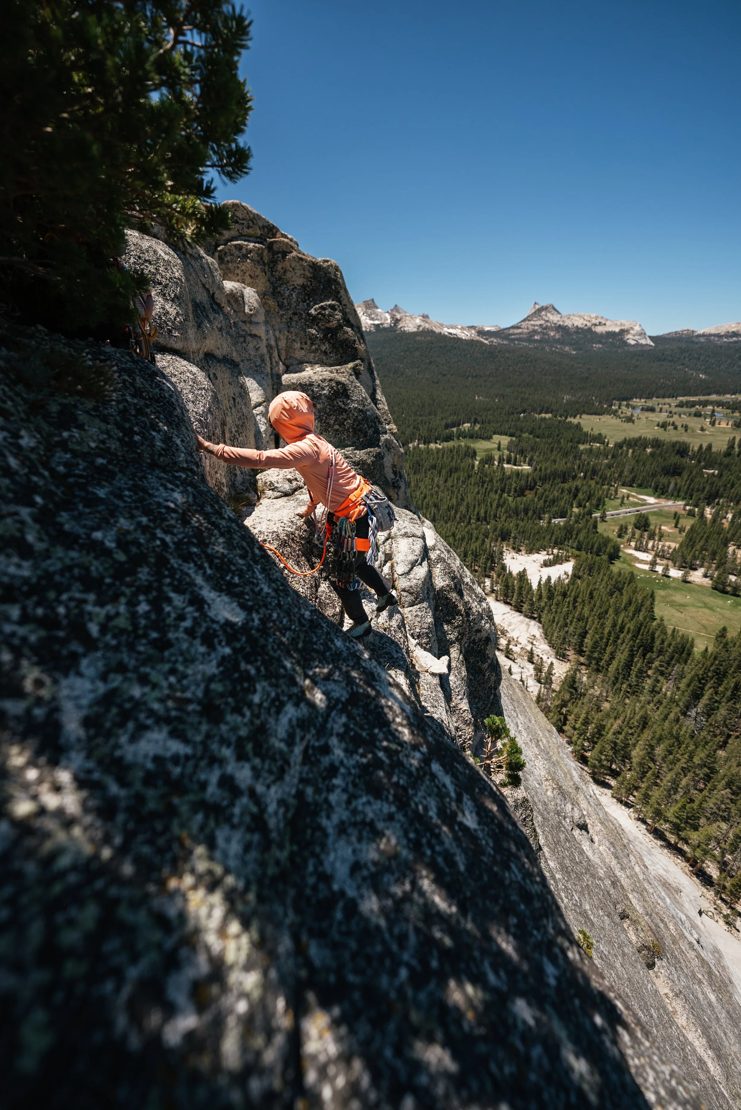 A person wearing climbing gear ascends a steep rock face with mountains and a forested valley in the background.