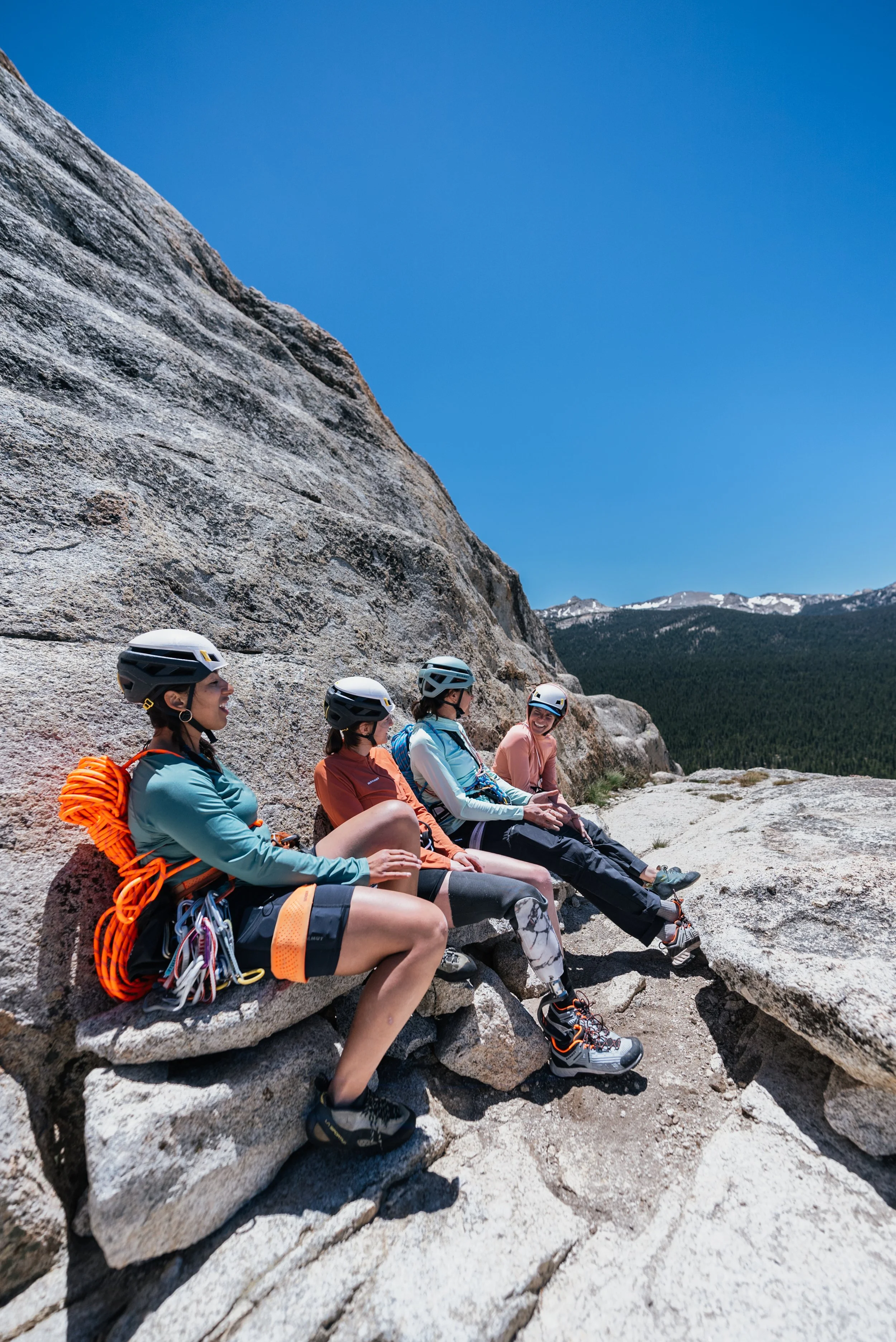 Four women wearing helmets and outdoor gear sitting on rocks against a mountain and clear blue sky.