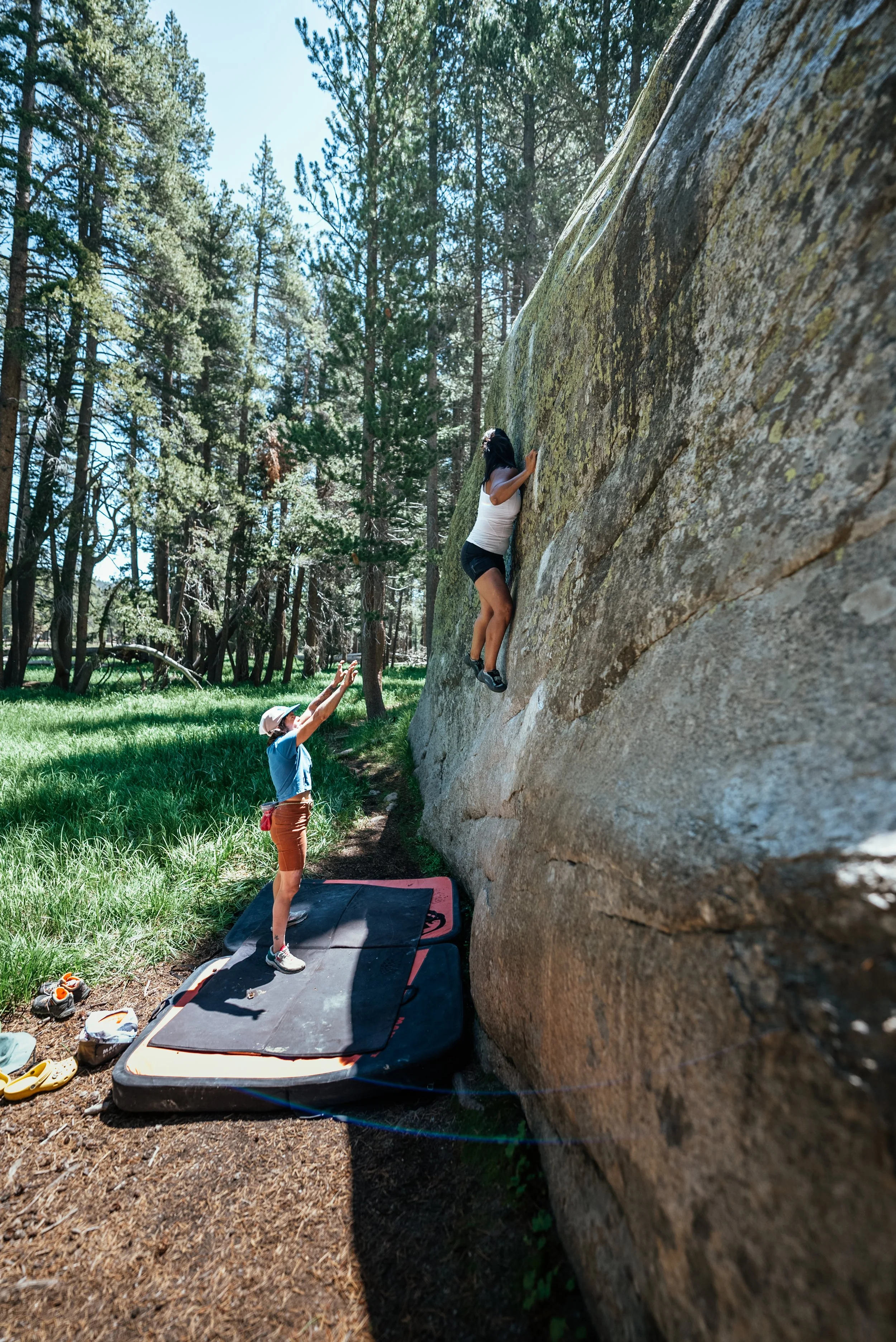 A woman is bouldering on an outdoor rock wall with a spotter below her in a forest setting. The spotter is reaching up to assist. Climbing shoes and gear are on the ground nearby.