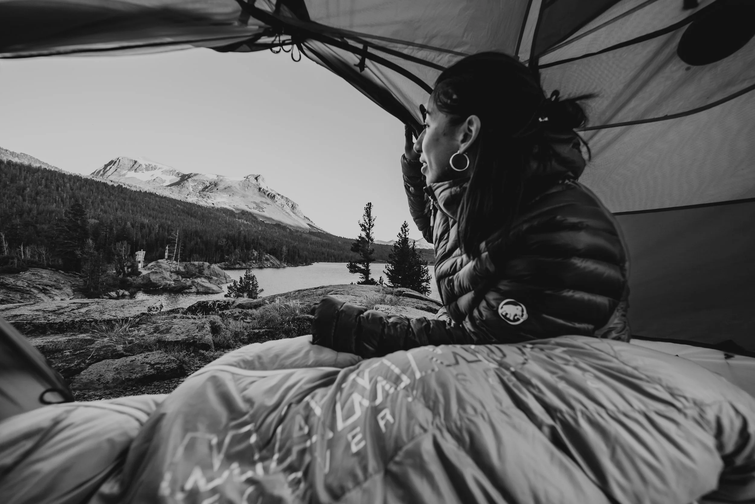 A woman sitting inside a camping tent overlooking a lake, with mountains and trees in the background.