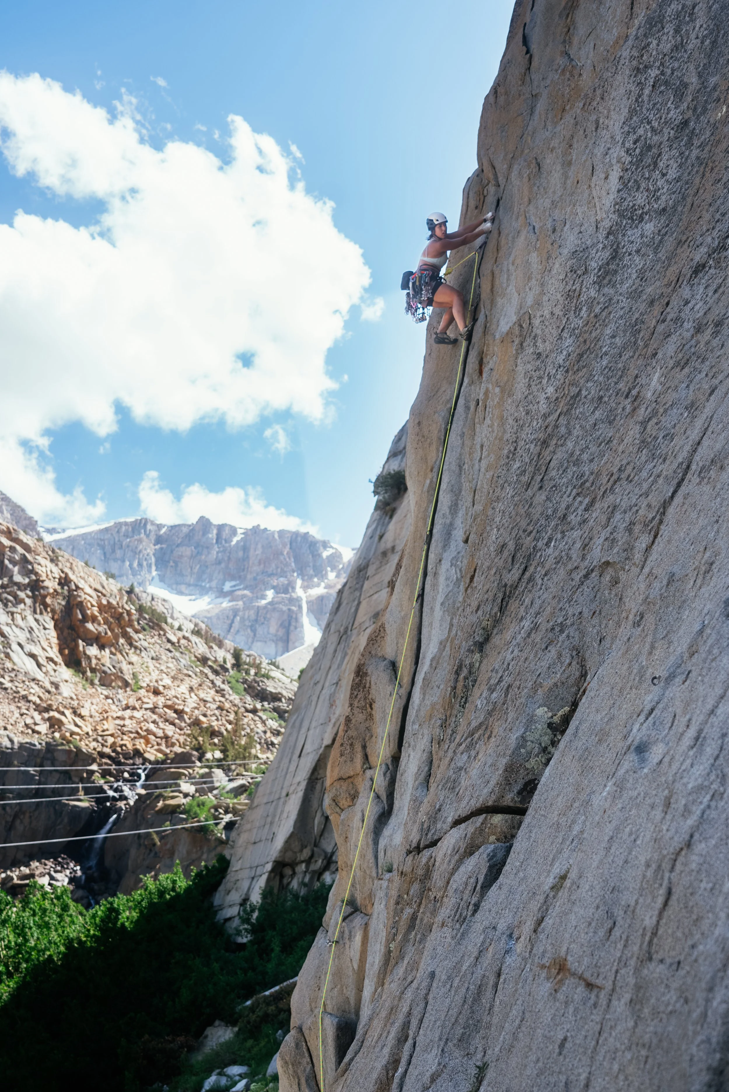A person rock climbing on a steep cliff face outdoors under a blue sky with clouds, mountains in the background, wearing a helmet and climbing gear.