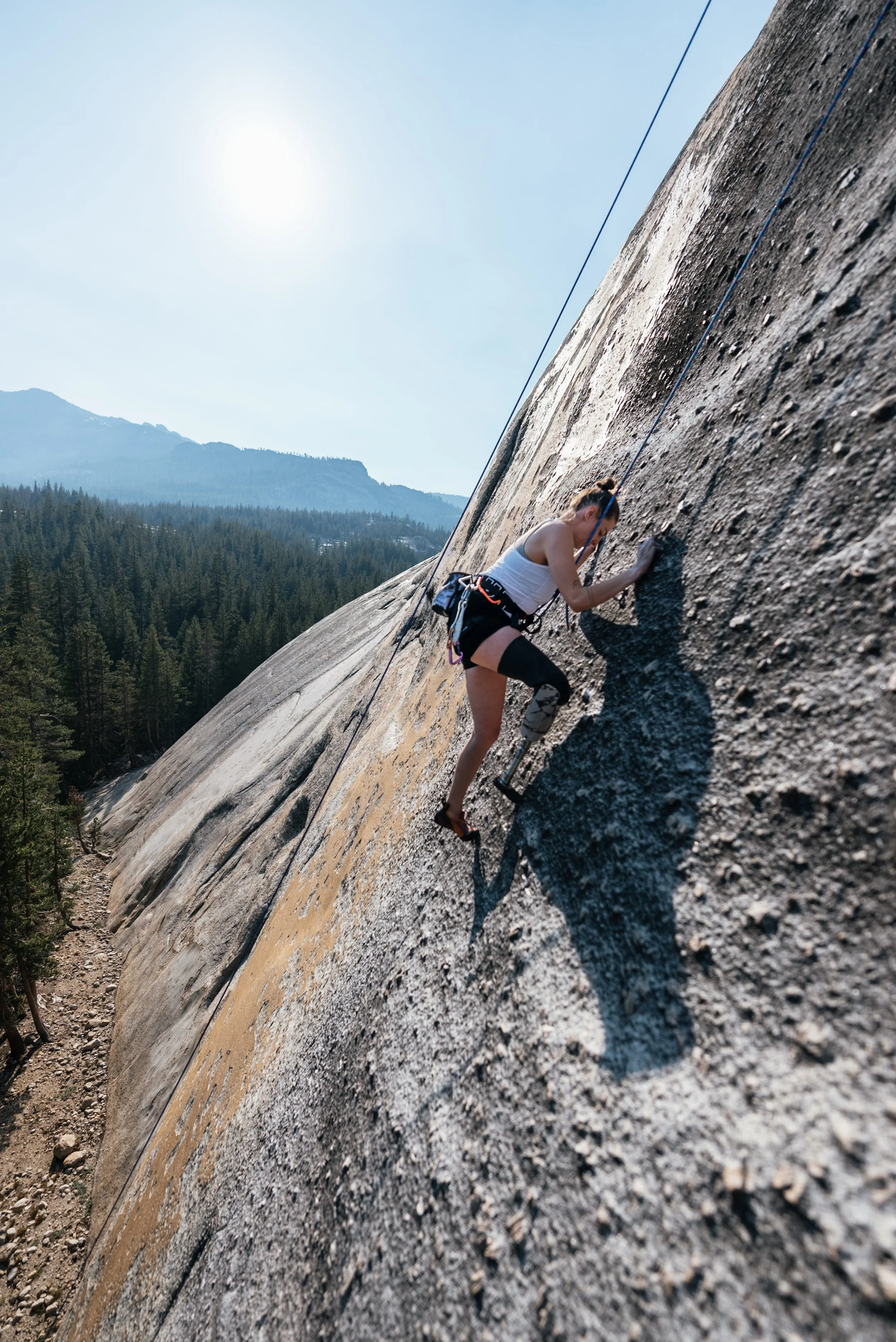 A woman rock climbing on a steep outdoor rock face with a scenic forest and mountains in the background, under a clear sky.