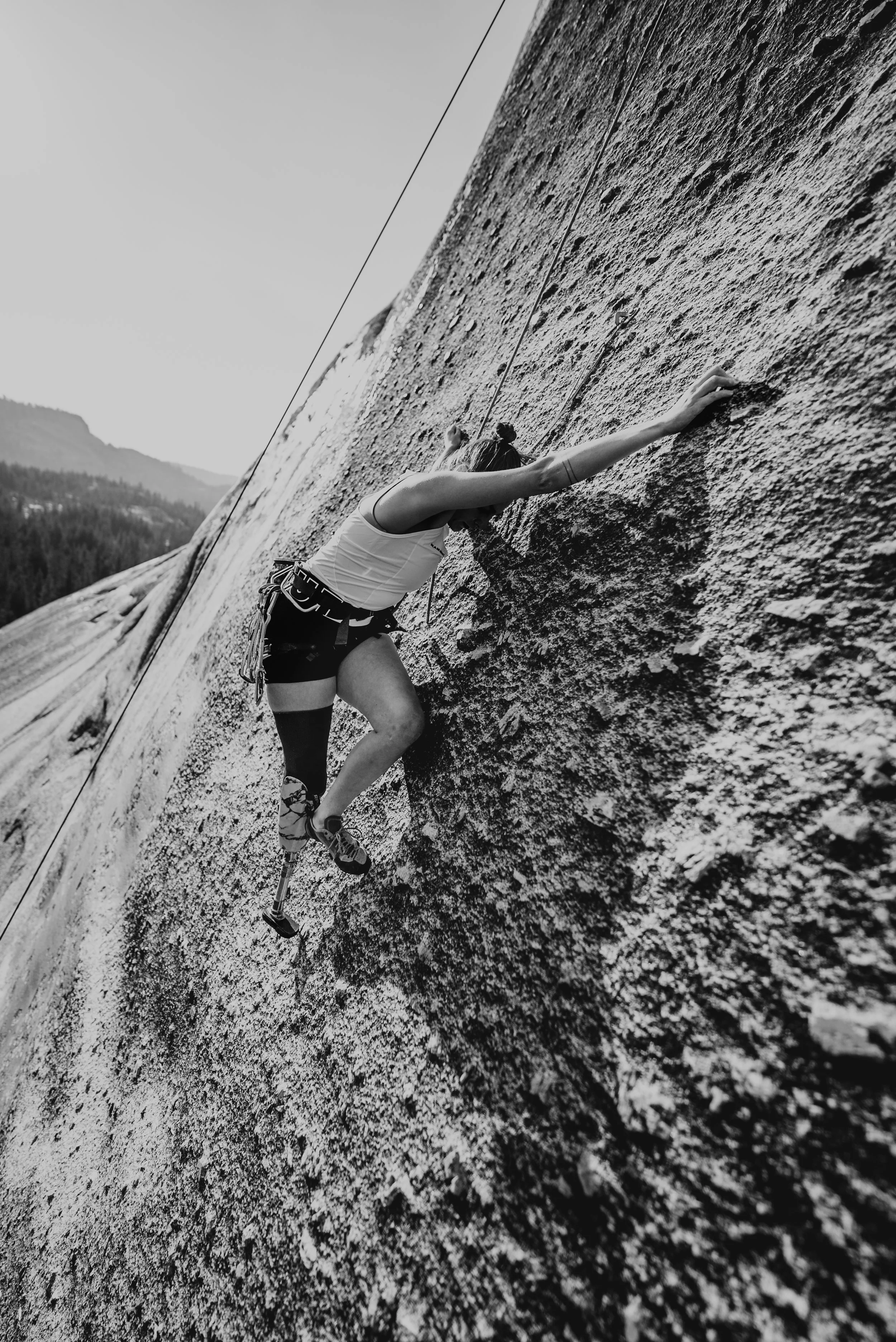 A person with a prosthetic leg rock climbing on a steep outdoor granite wall, wearing climbing gear and a tank top.