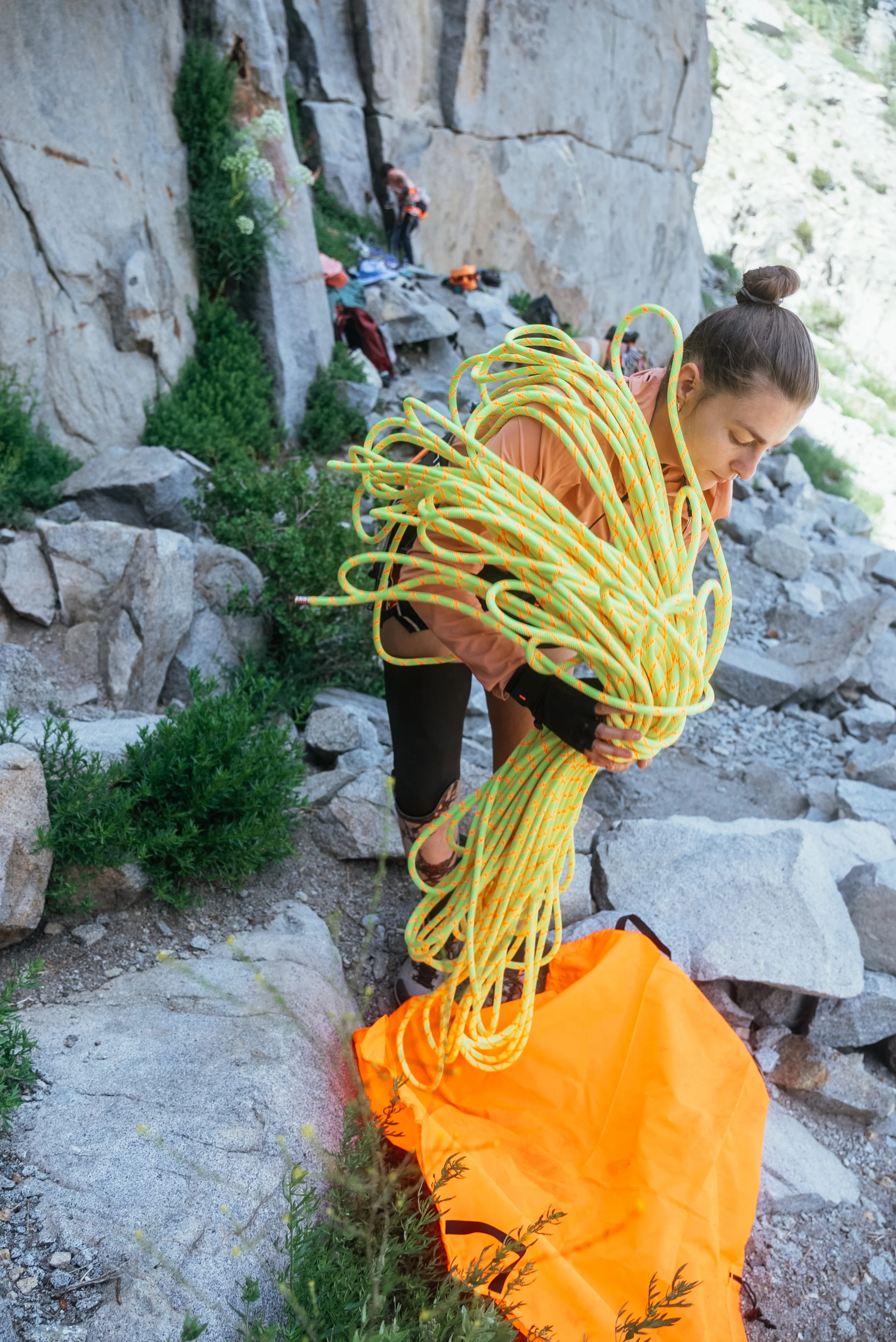 A woman on a rocky mountain trail carrying a large bundle of climbing ropes next to an orange gear bag with shrubbery and other climbers in the background.