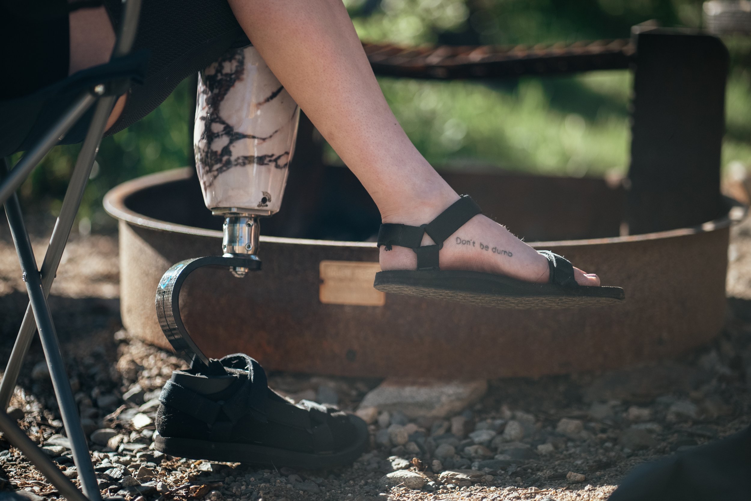 A person with a prosthetic leg sitting on a folding camp chair, wearing black sandals, with a tattoo that says "Don't be dumb" on their ankle, near a fire pit outdoors.