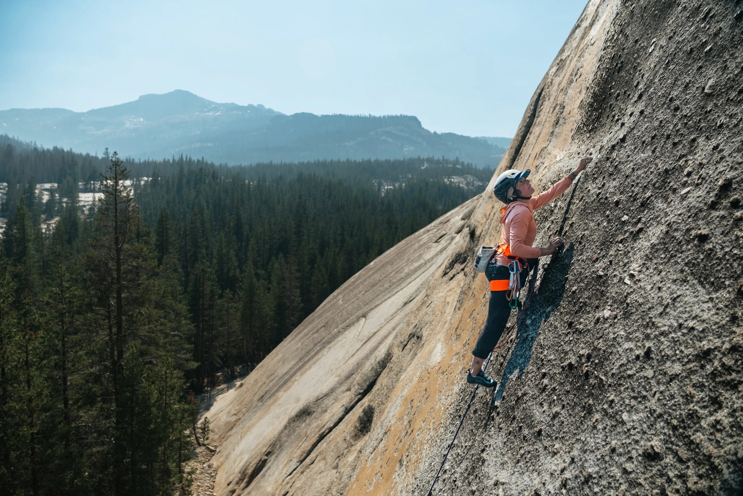 A woman rock climbing on a granite cliff face, wearing a helmet and safety harness, with a scenic forested mountain landscape in the background.