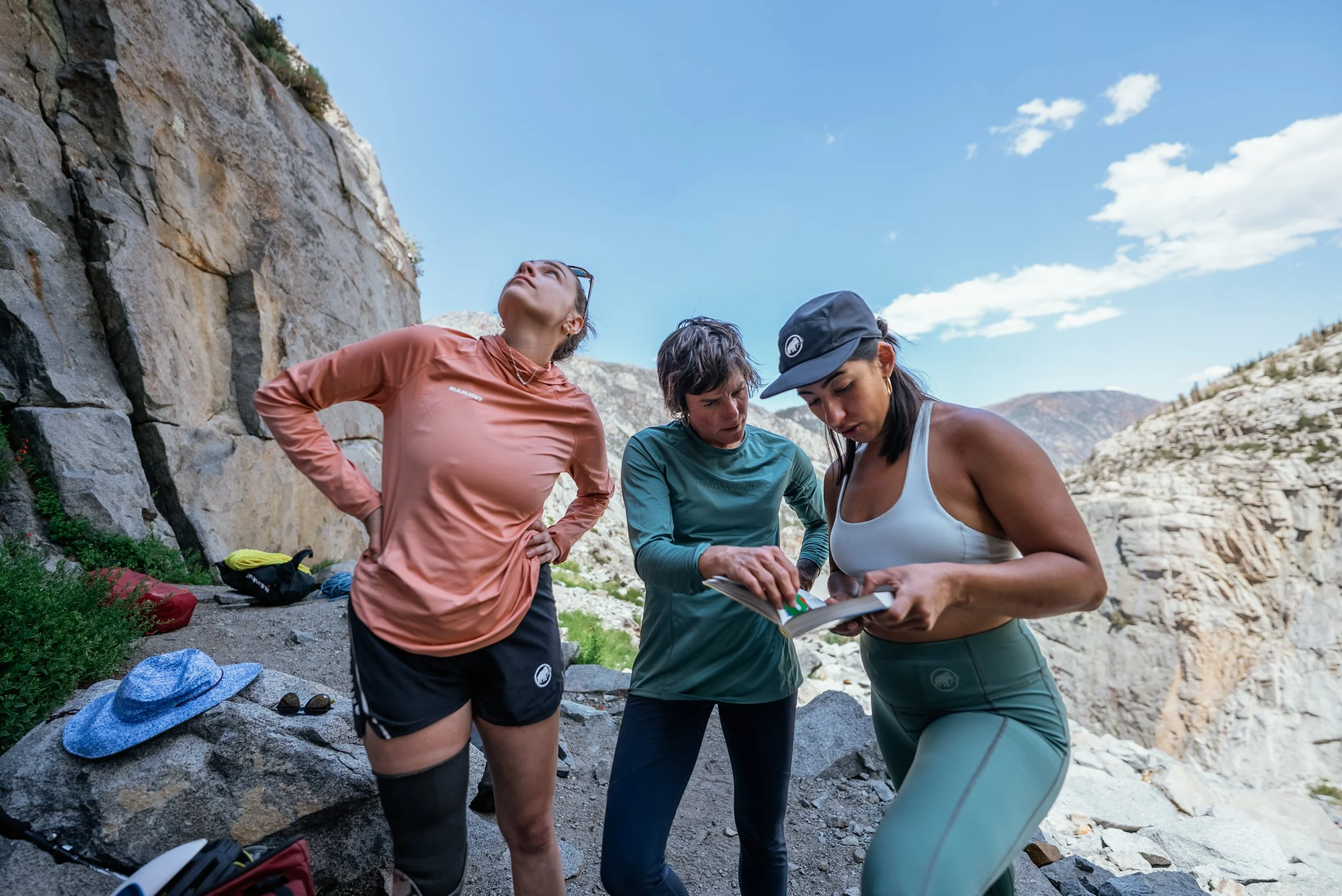 Three women on a mountain hike examining a map or guidebook, surrounded by rocky terrain and clear blue sky.