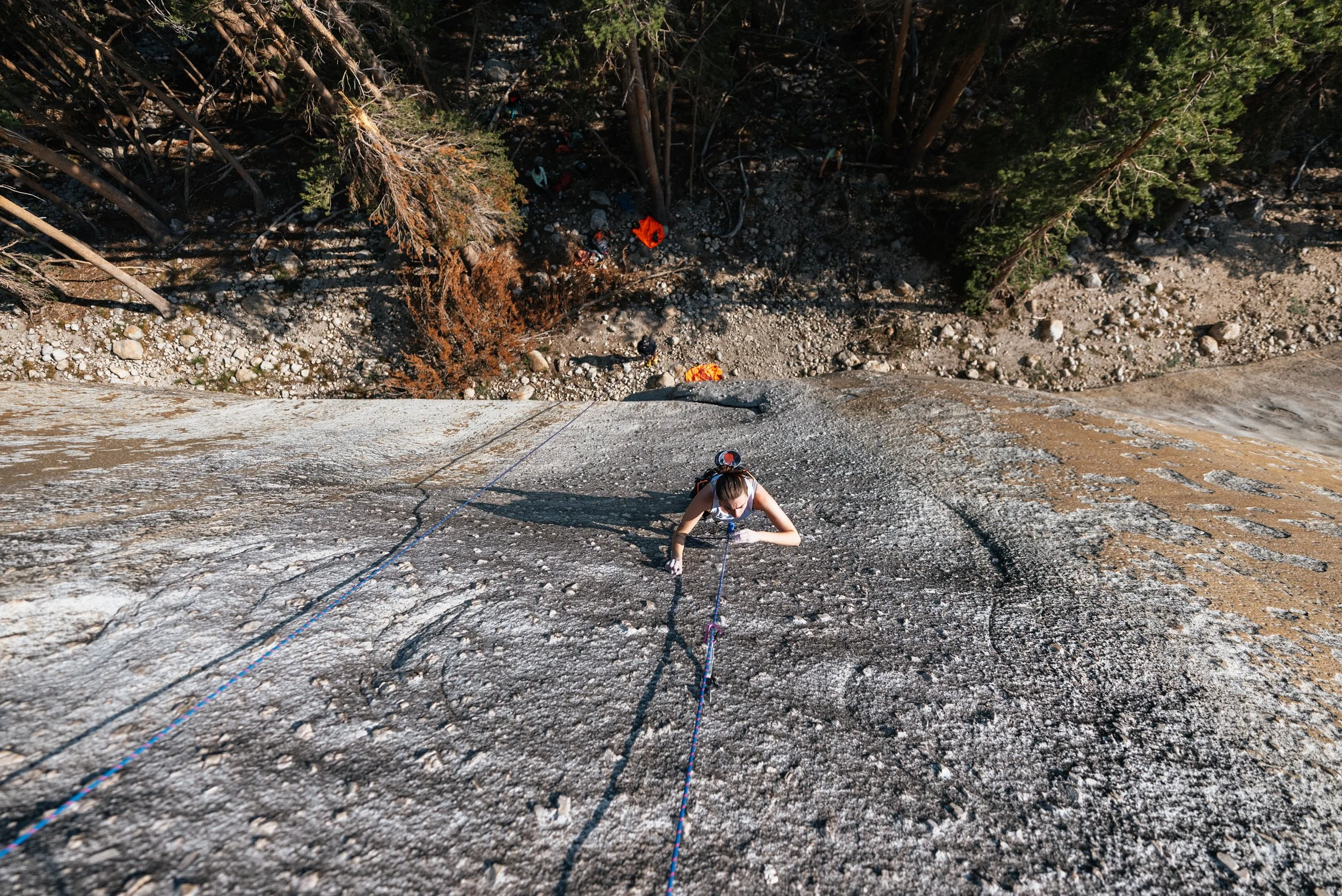 A person rock climbing on a steep outdoor rock face, viewed from above, with trees and a rocky ground visible below.