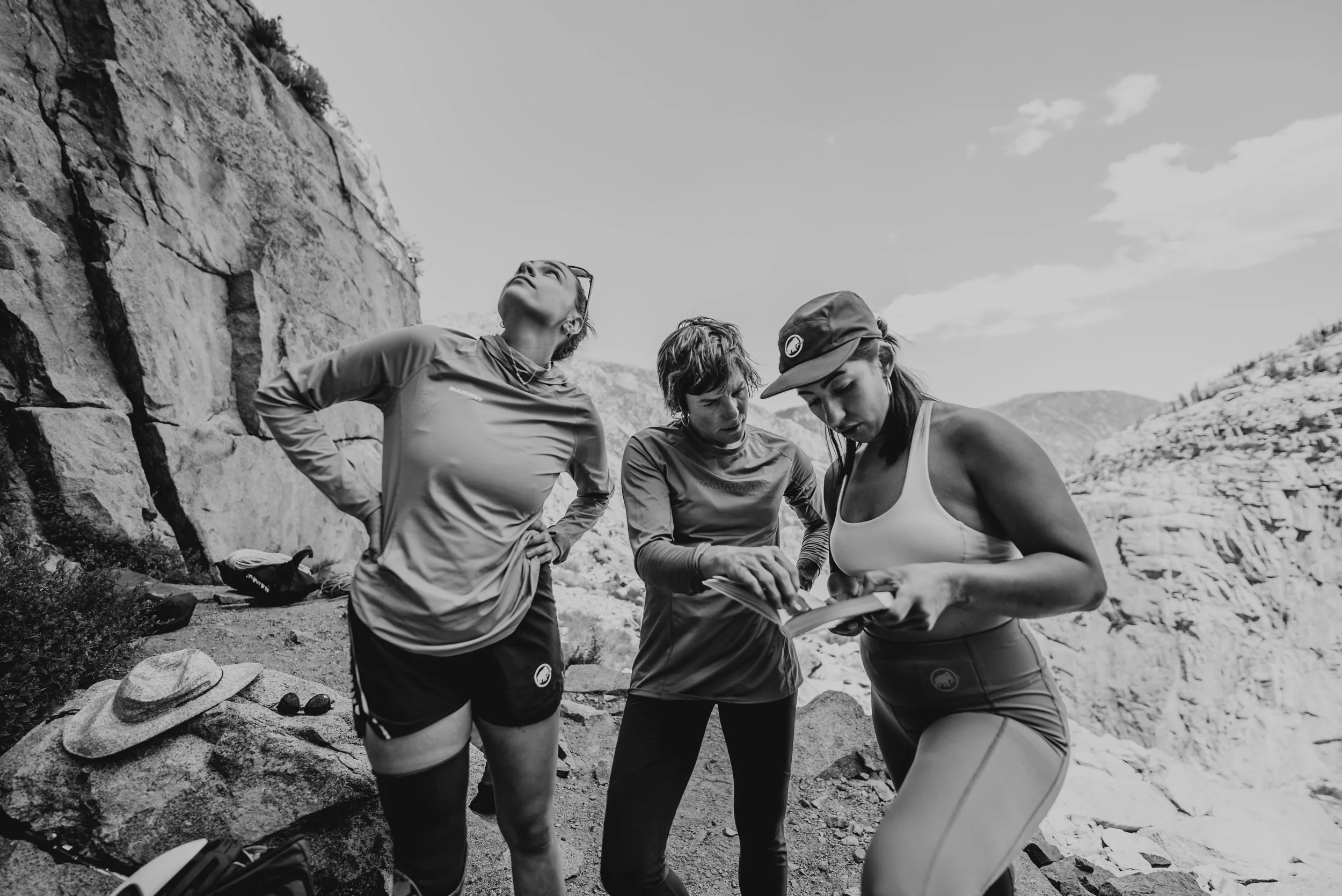 Three women hikers outdoors near rocky cliffs, with one showing something on a notebook, in black and white.