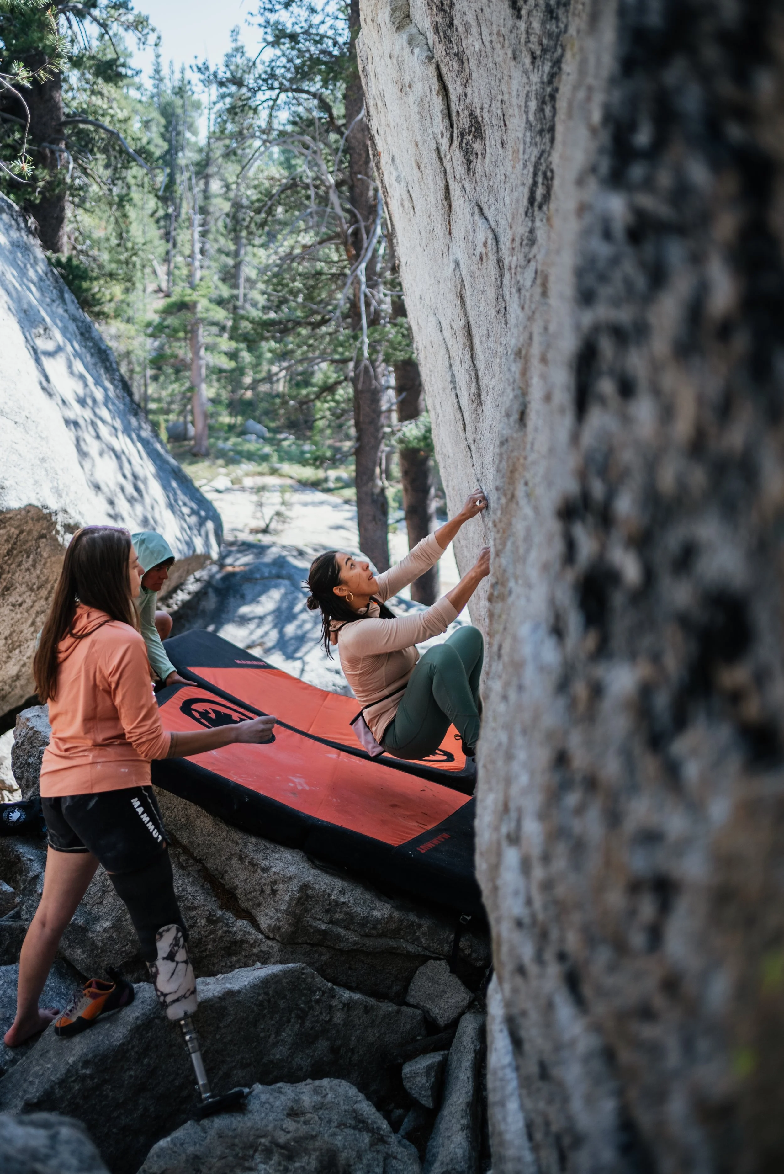 Three women at a bouldering site outdoors, two helping a third woman who is climbing a rock wall.