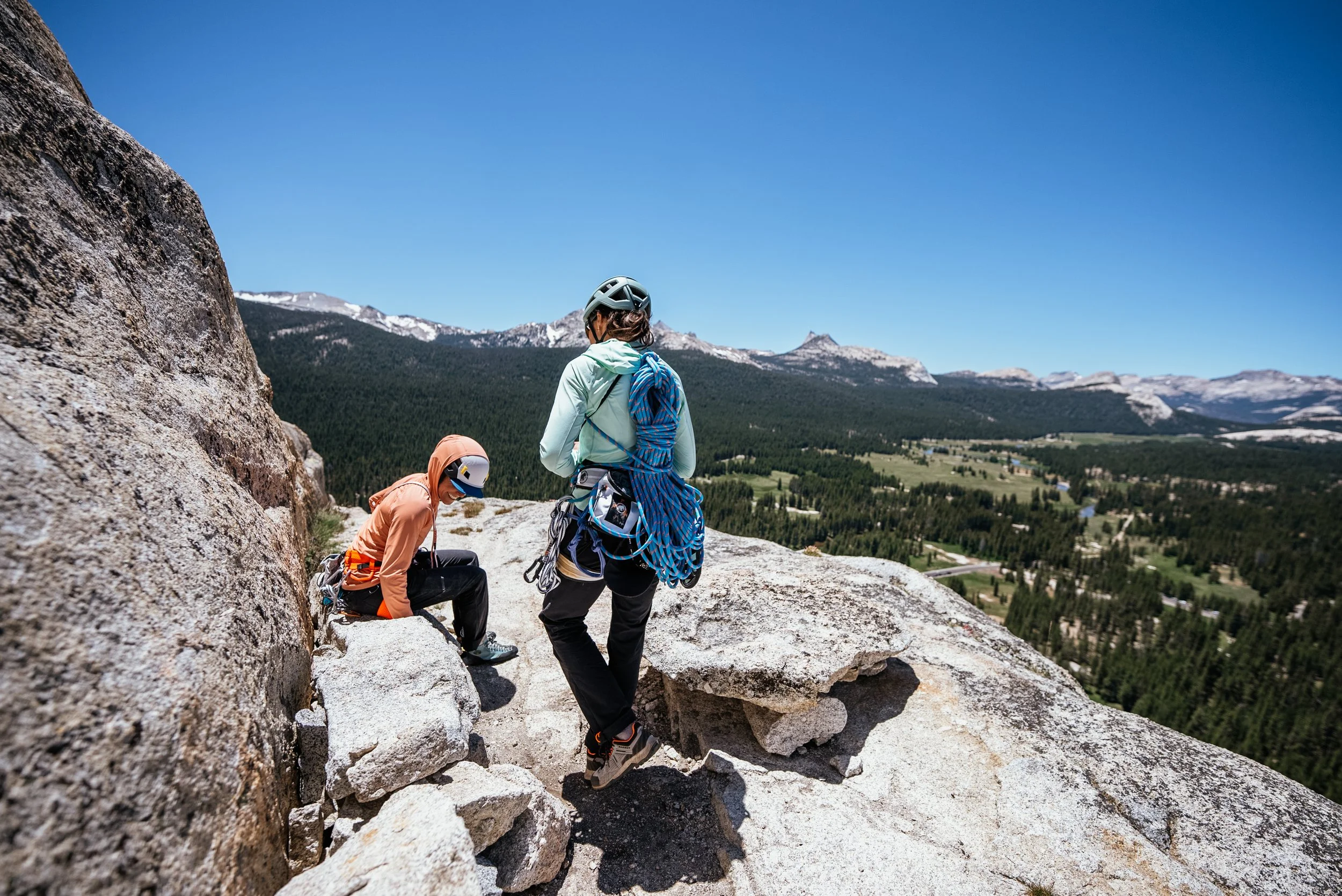 Two climbers on a rocky mountain ledge overlooking a valley with forests, rivers, and mountains under a clear blue sky.