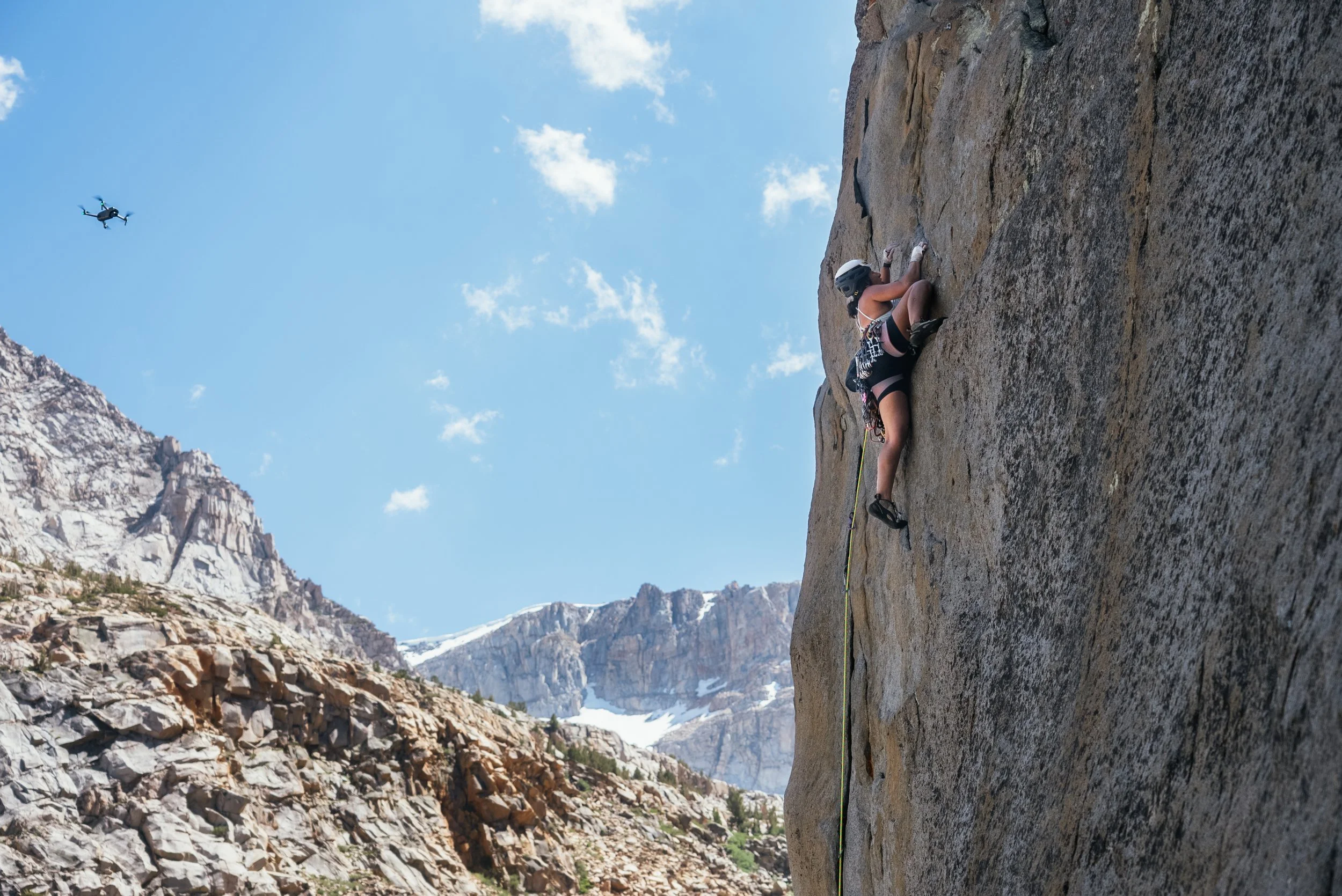 A woman rock climbing on a vertical cliff face with mountains and a blue sky in the background, and a drone flying nearby.