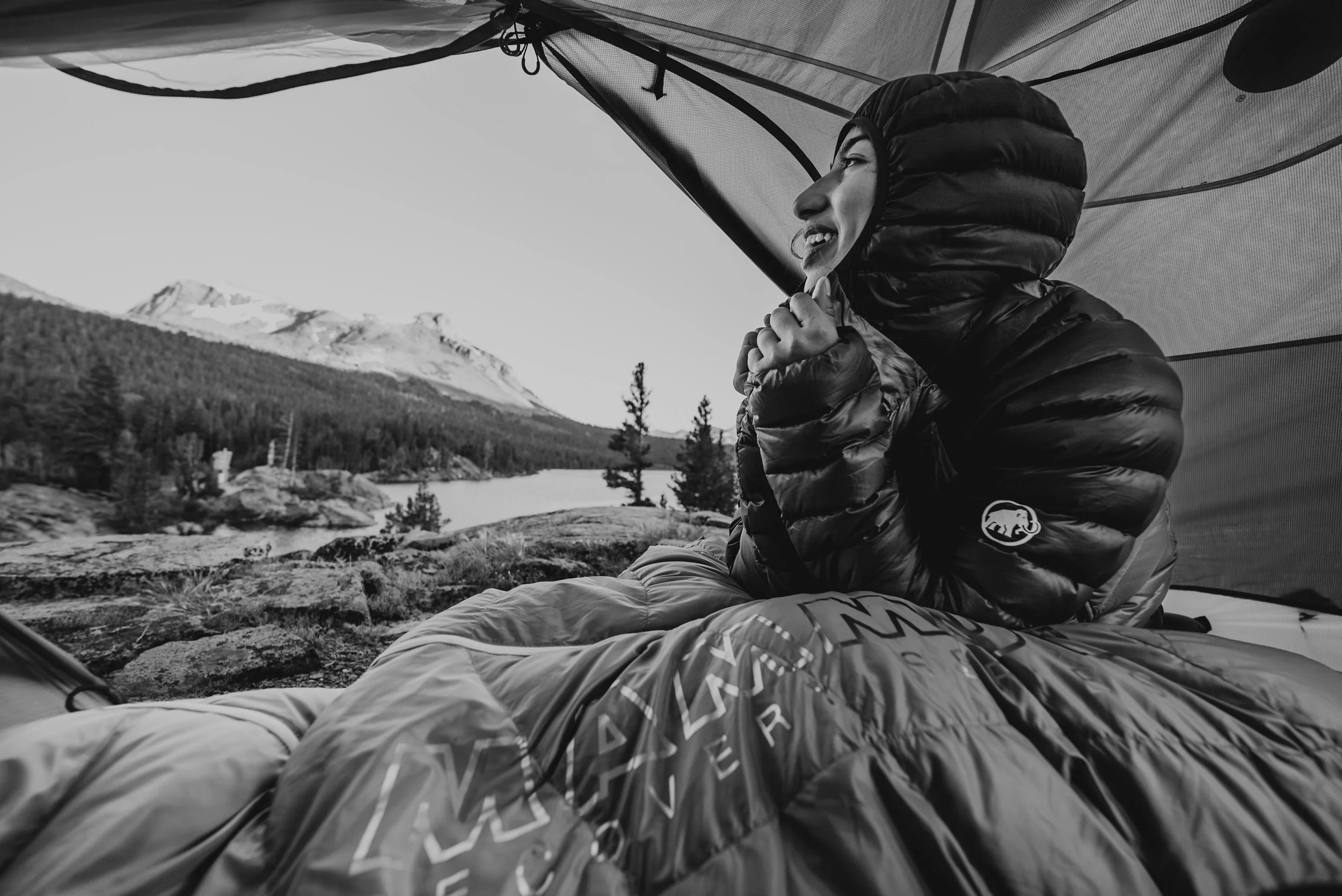 A woman sitting inside a tent on a rocky terrain, wearing a puffy jacket with a hood, smiling and looking outside at a mountain and pine trees near a lake.