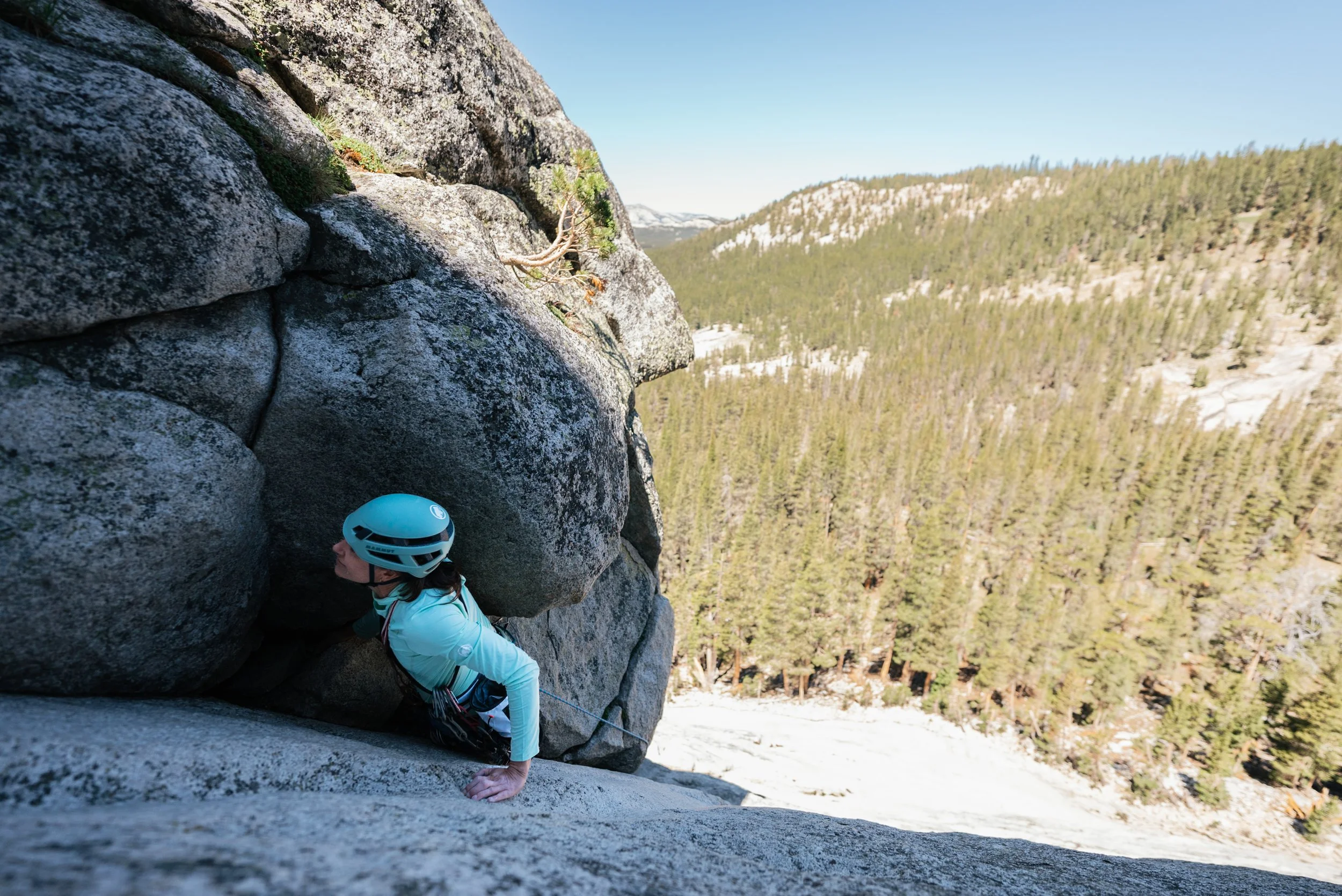 A woman climbing a rock face in a mountainous forested area with pine trees and snow, wearing a blue helmet and climbing gear.