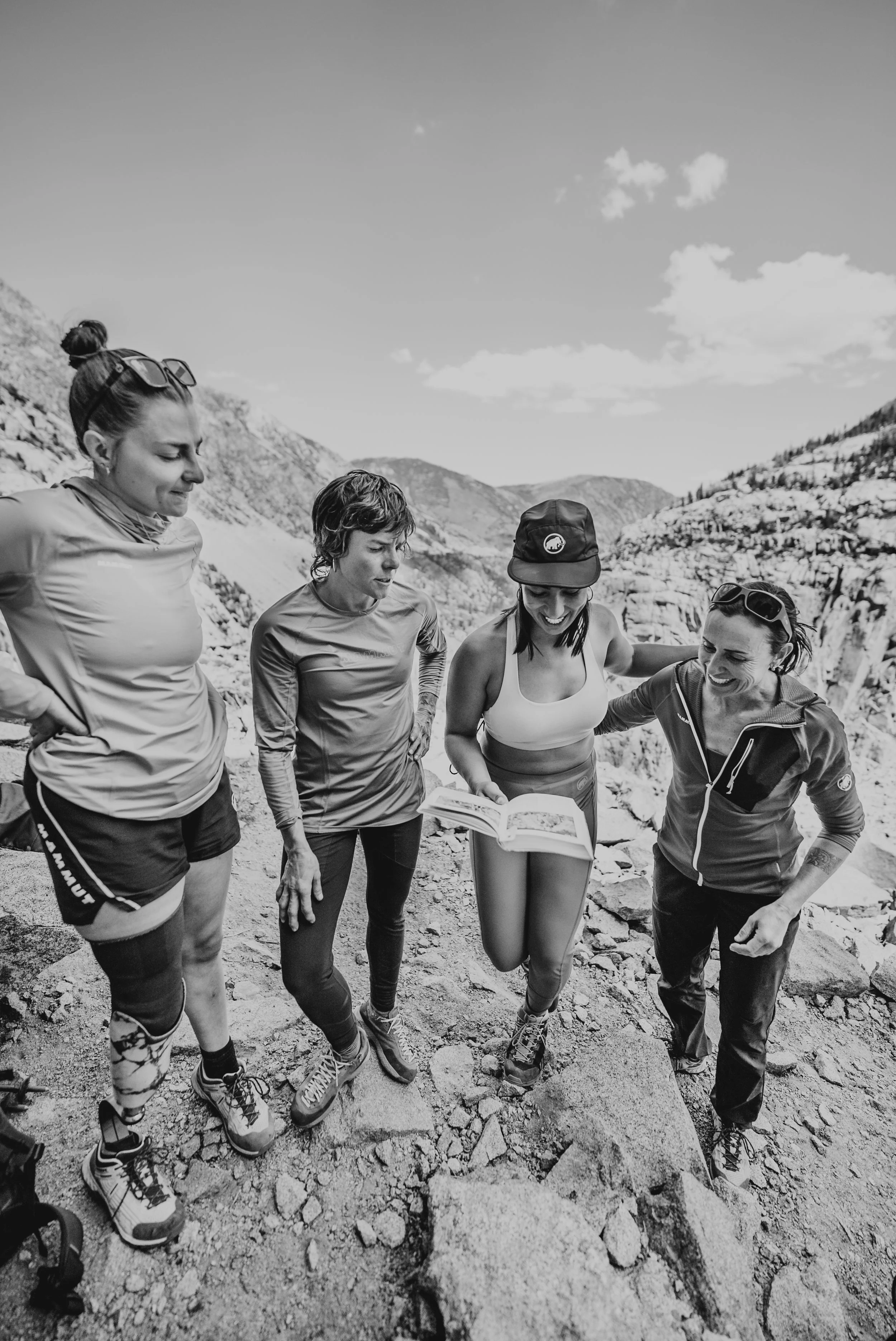 Four women in outdoor athletic clothing standing on rocky terrain, looking at a map, surrounded by mountainous landscape.