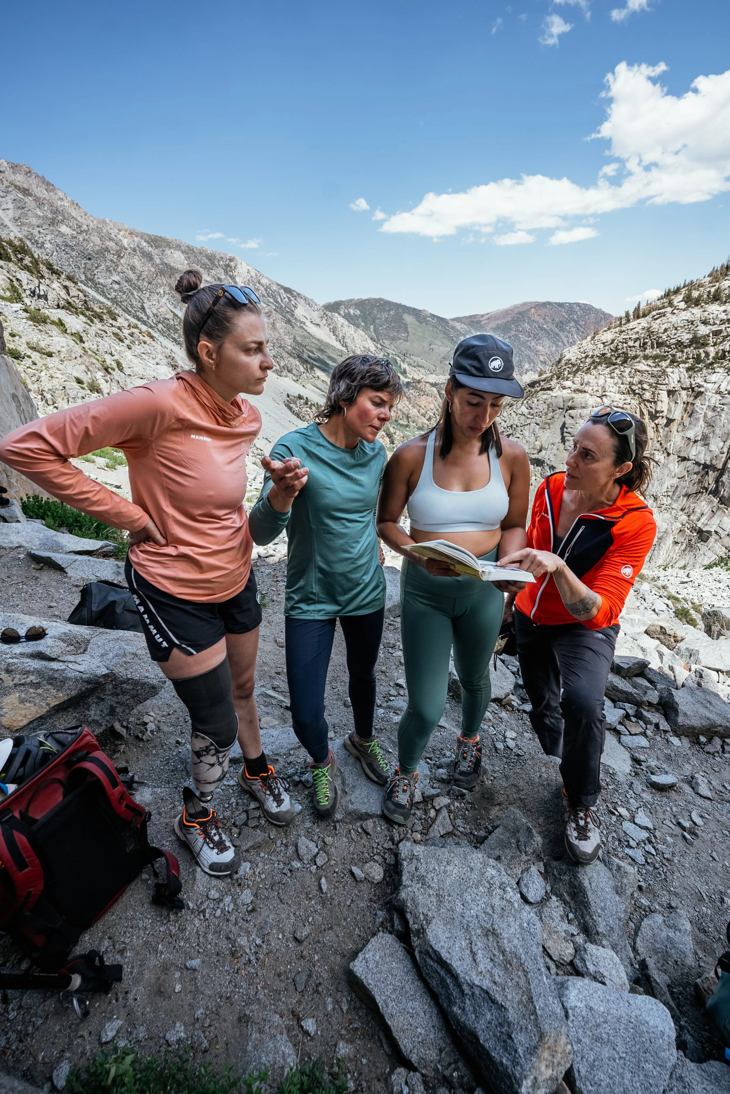 Four women hikers looking at a map together on a rocky mountain trail with rugged hills and blue sky in the background.