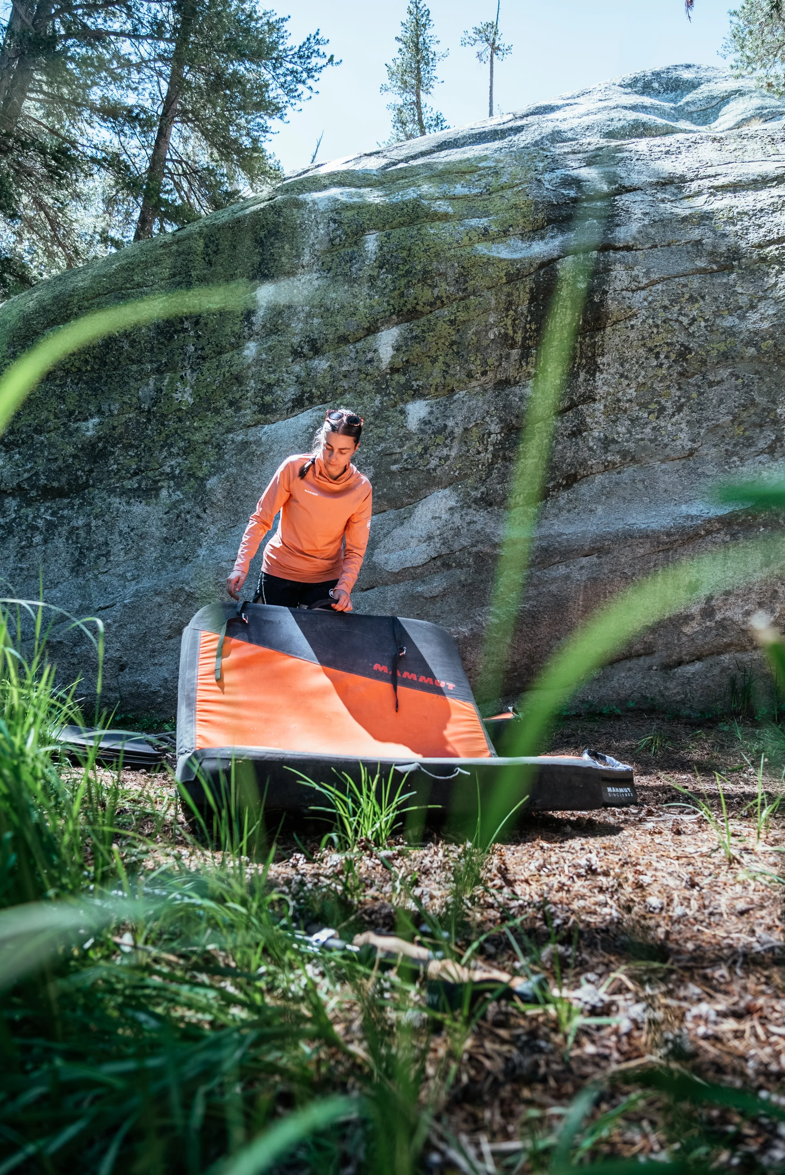 A woman in an orange jacket unfolding or preparing a Mammut avalanche airbag backpack in a natural outdoor setting with large rock formations and green trees in the background.