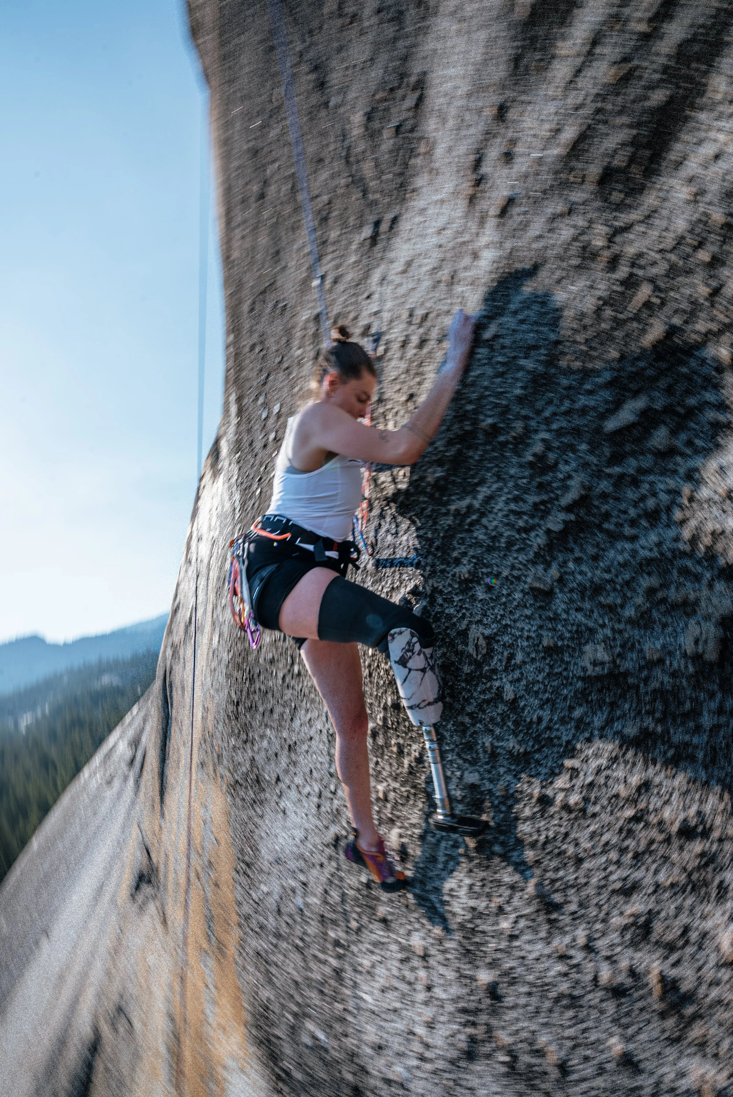 A woman with a prosthetic leg rock climbing a steep granite wall outdoors, with mountains and trees in the background.