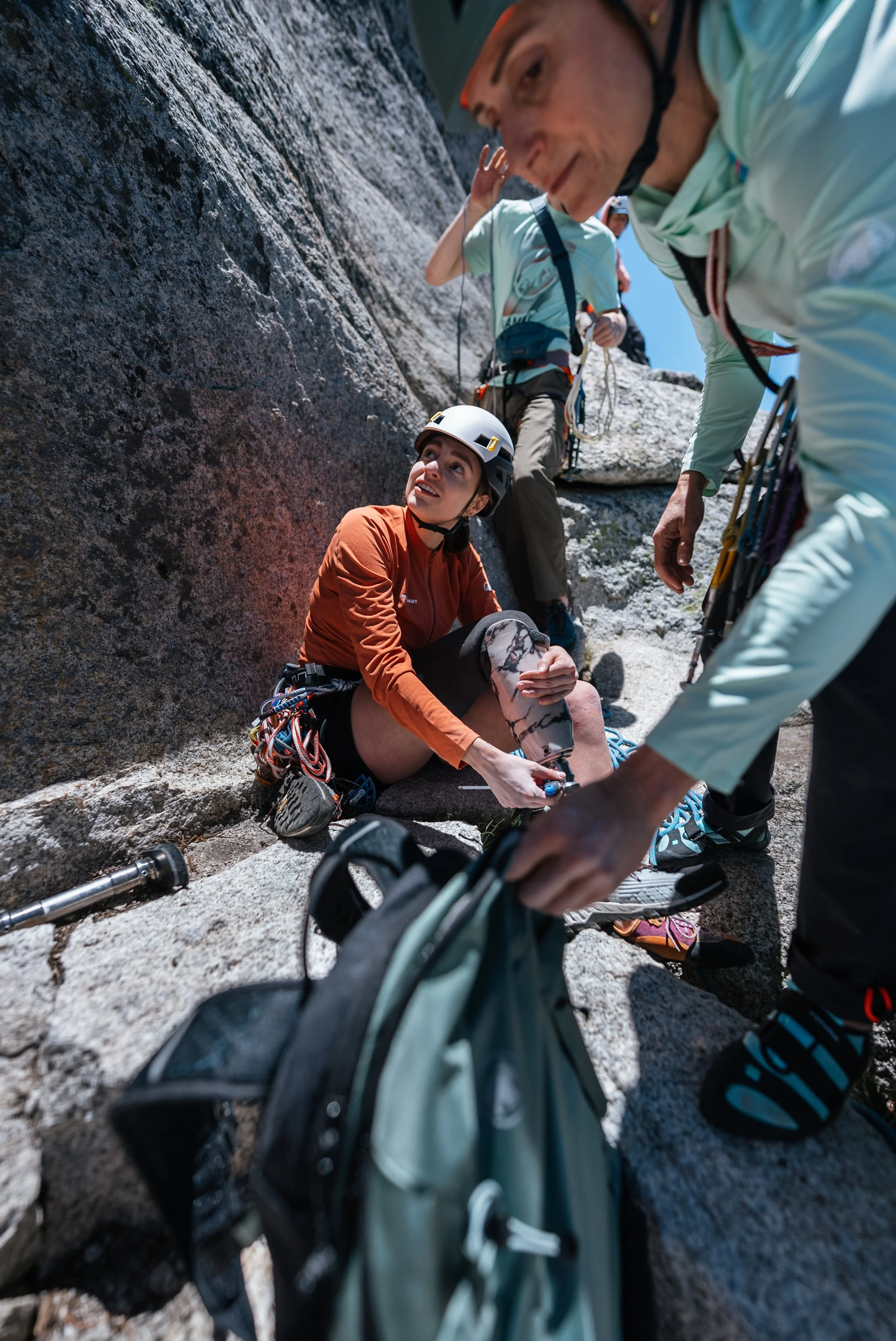A female rock climber with a helmet sitting on the ground and receiving care from her climbing partners on a rocky ledge during a mountain climb.