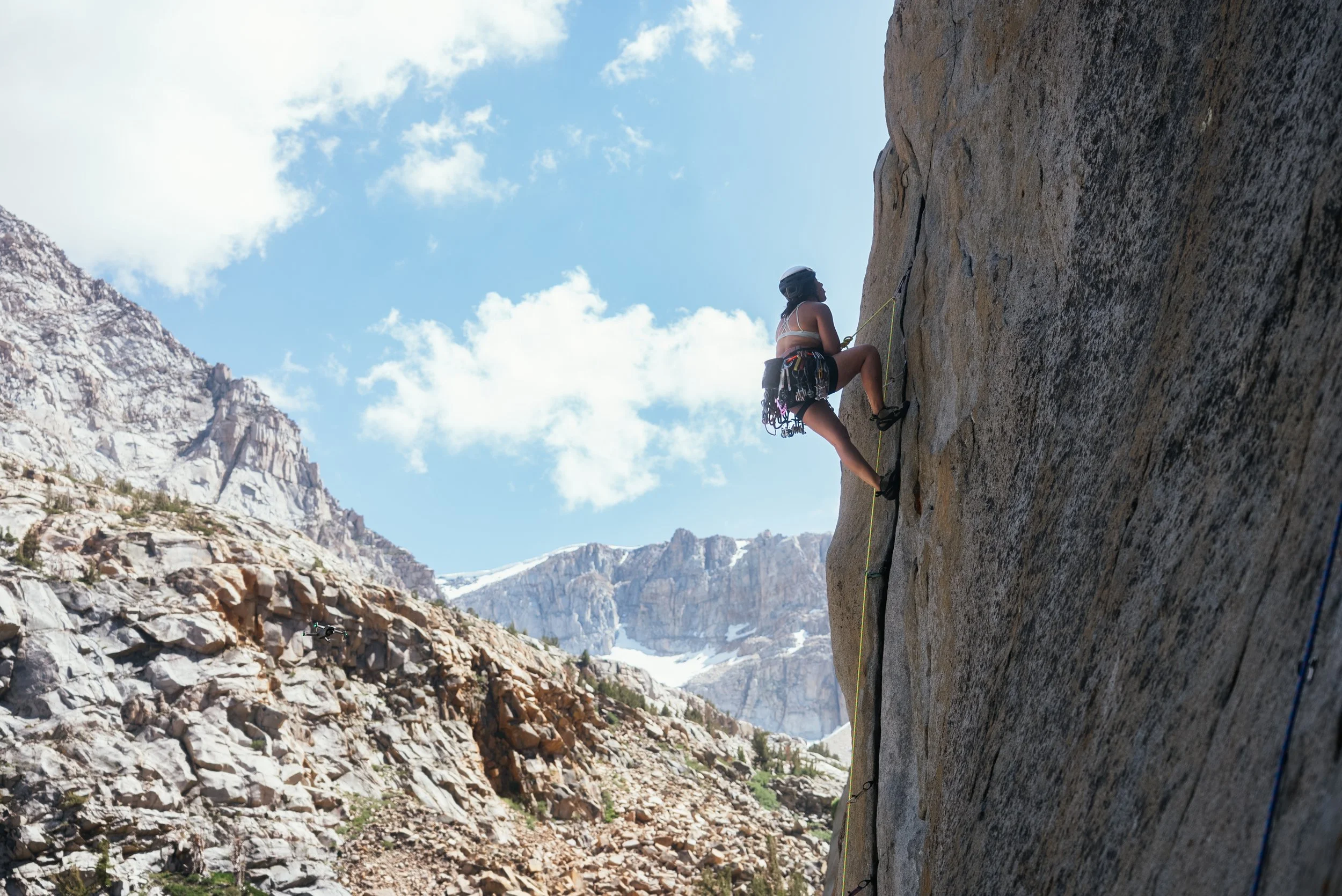 Woman rock climbing on a steep cliff with mountains in the background under a partly cloudy sky.