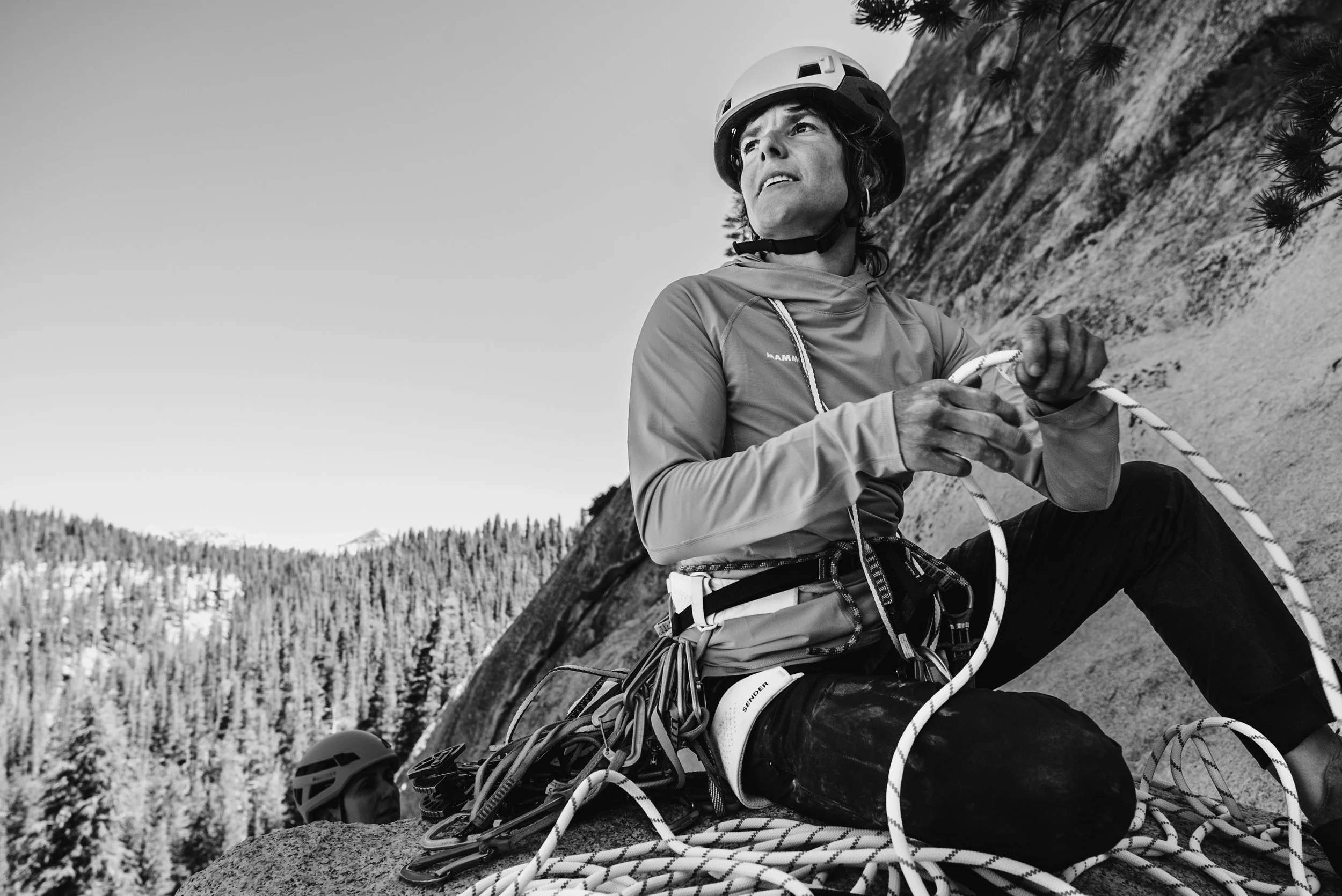 A person wearing a helmet and climbing gear, sitting on a rock, preparing for rock climbing in a mountainous area with trees in the background.