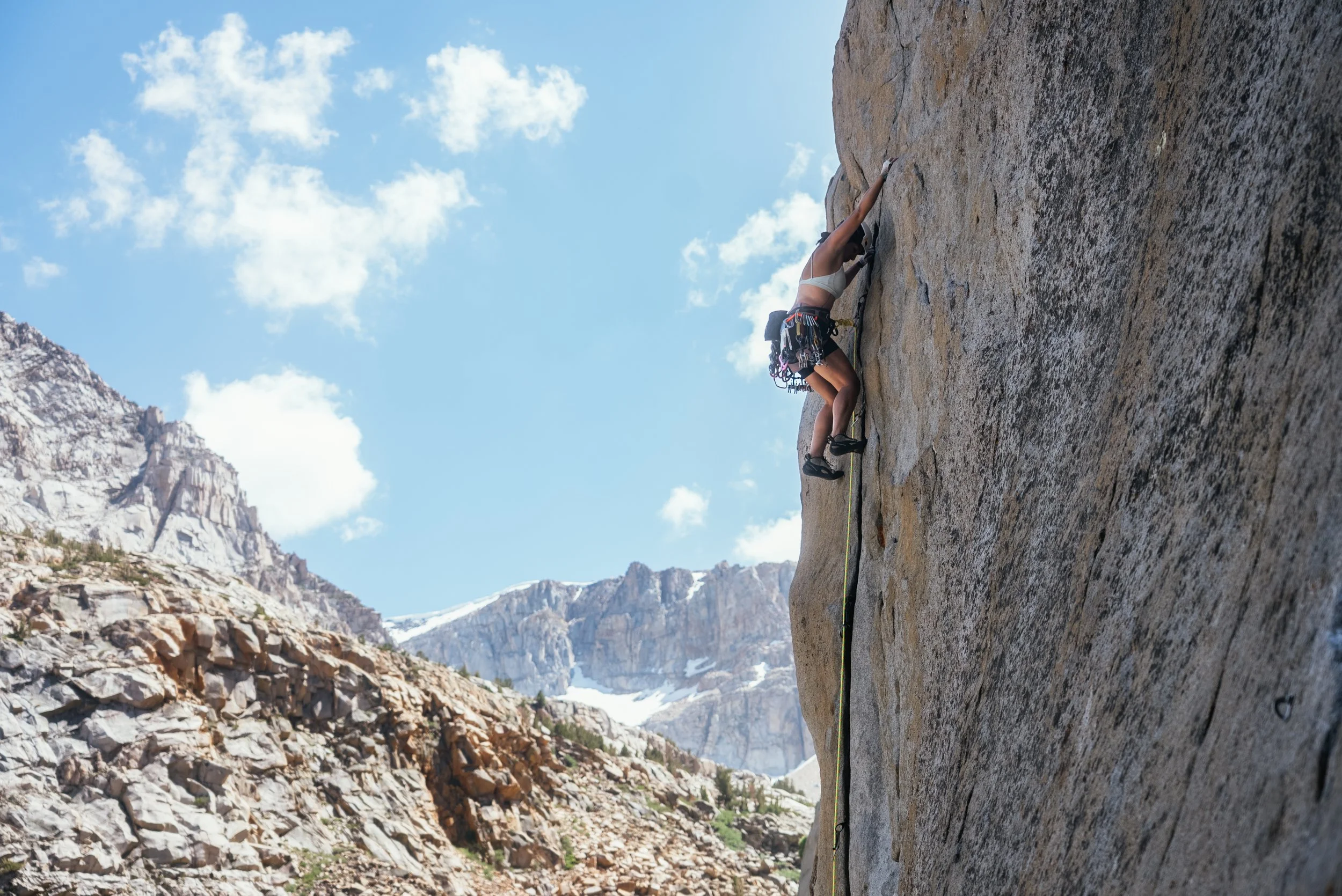 A woman rock climbing on a steep cliff face with mountains and a blue sky in the background.