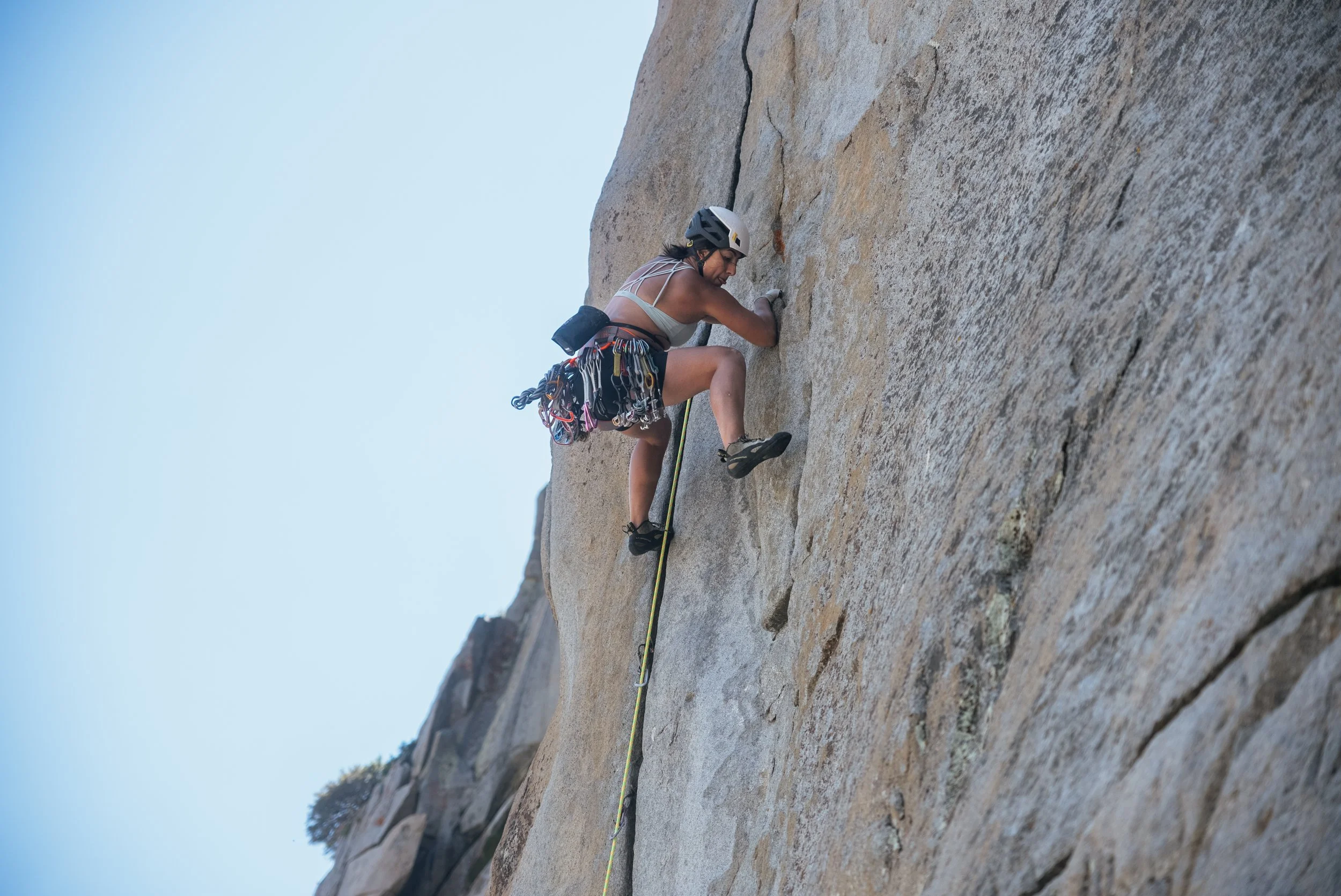 A woman climbing a steep rock face outdoors, wearing a helmet, climbing harness, and carrying climbing gear.