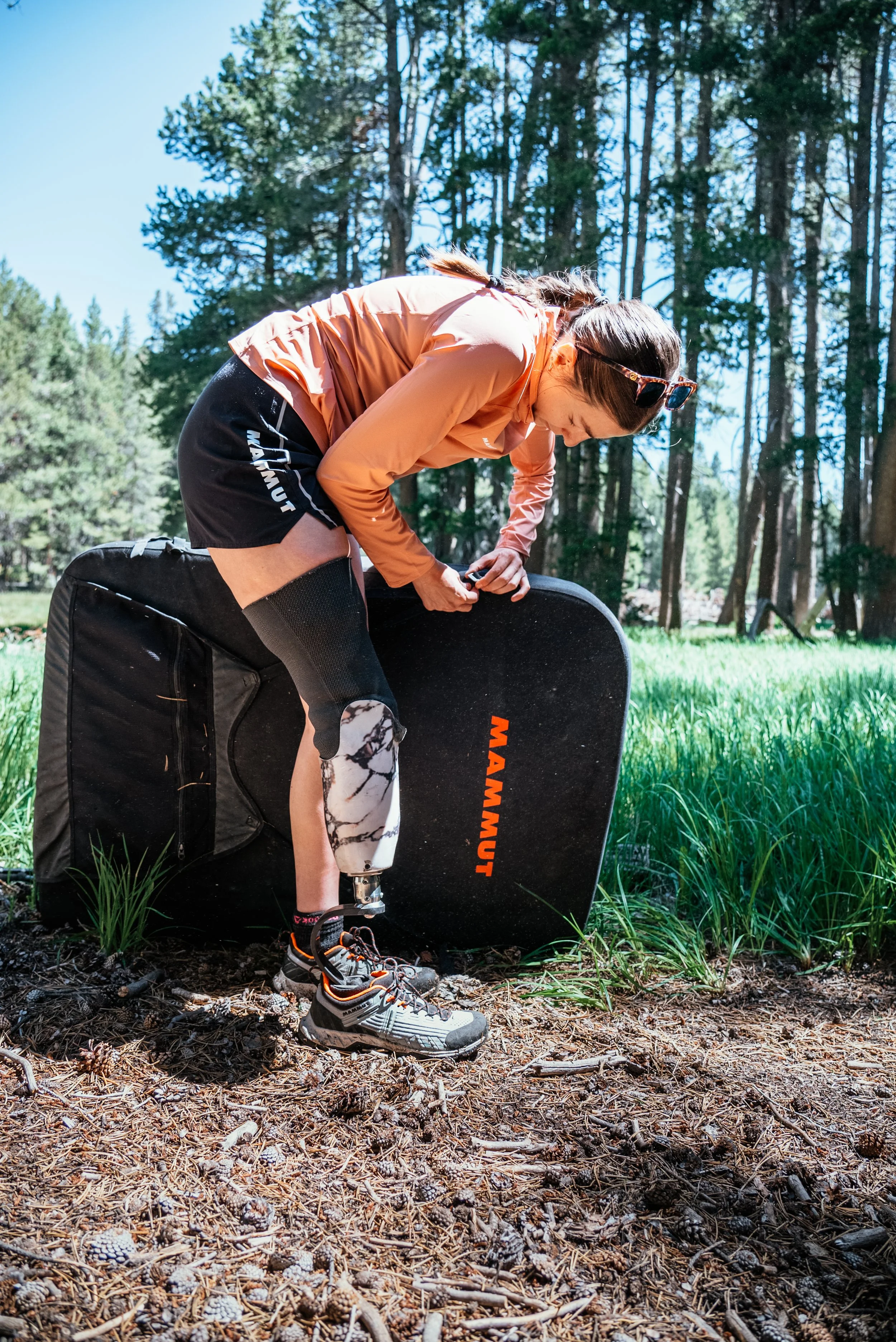 Woman in outdoor gear adjusting a Mamunt foamball in a forest setting with tall trees and green grass.