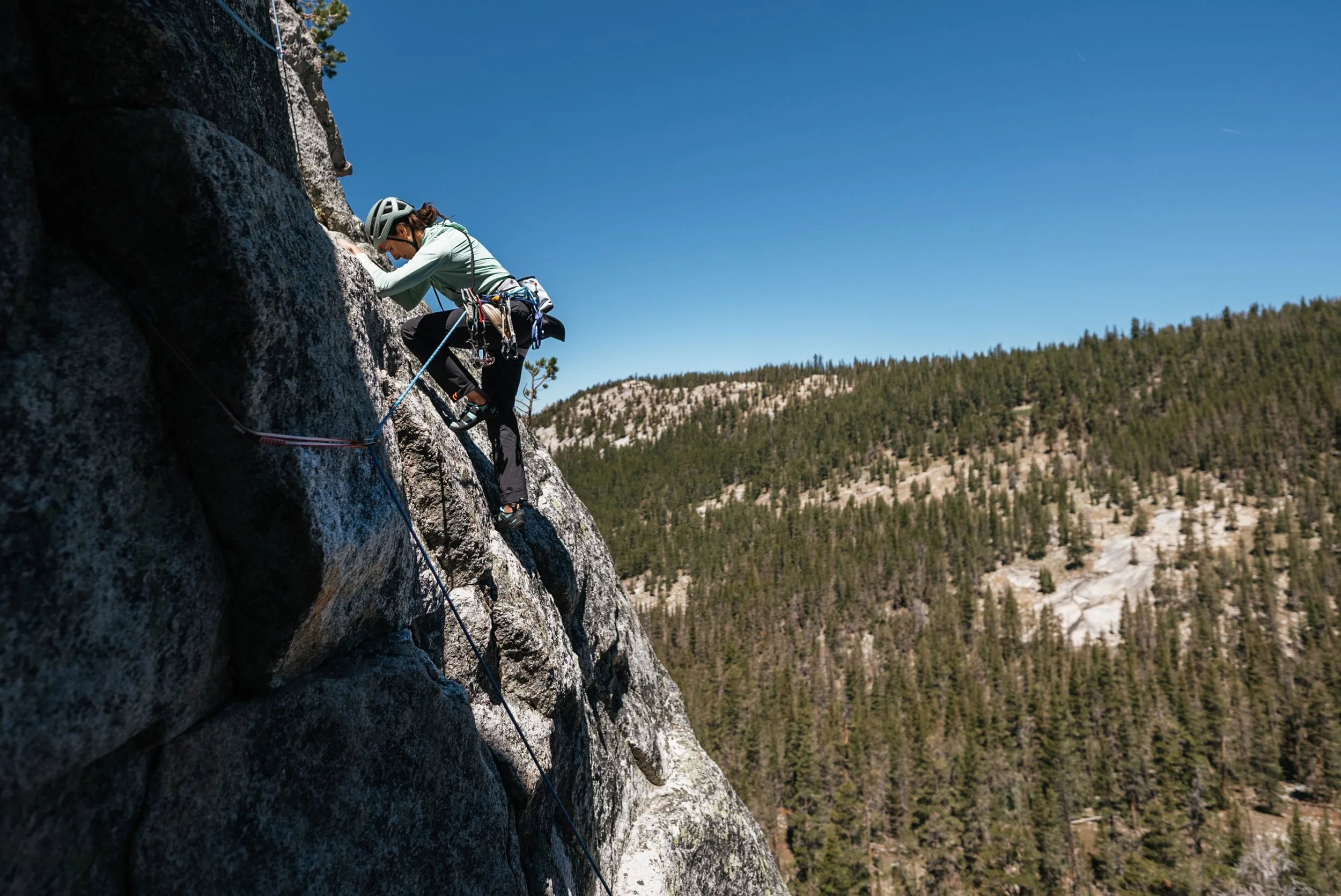 A person rock climbing on a steep granite cliff, wearing a helmet and climbing gear, with a landscape of trees and mountains in the background under a clear blue sky.