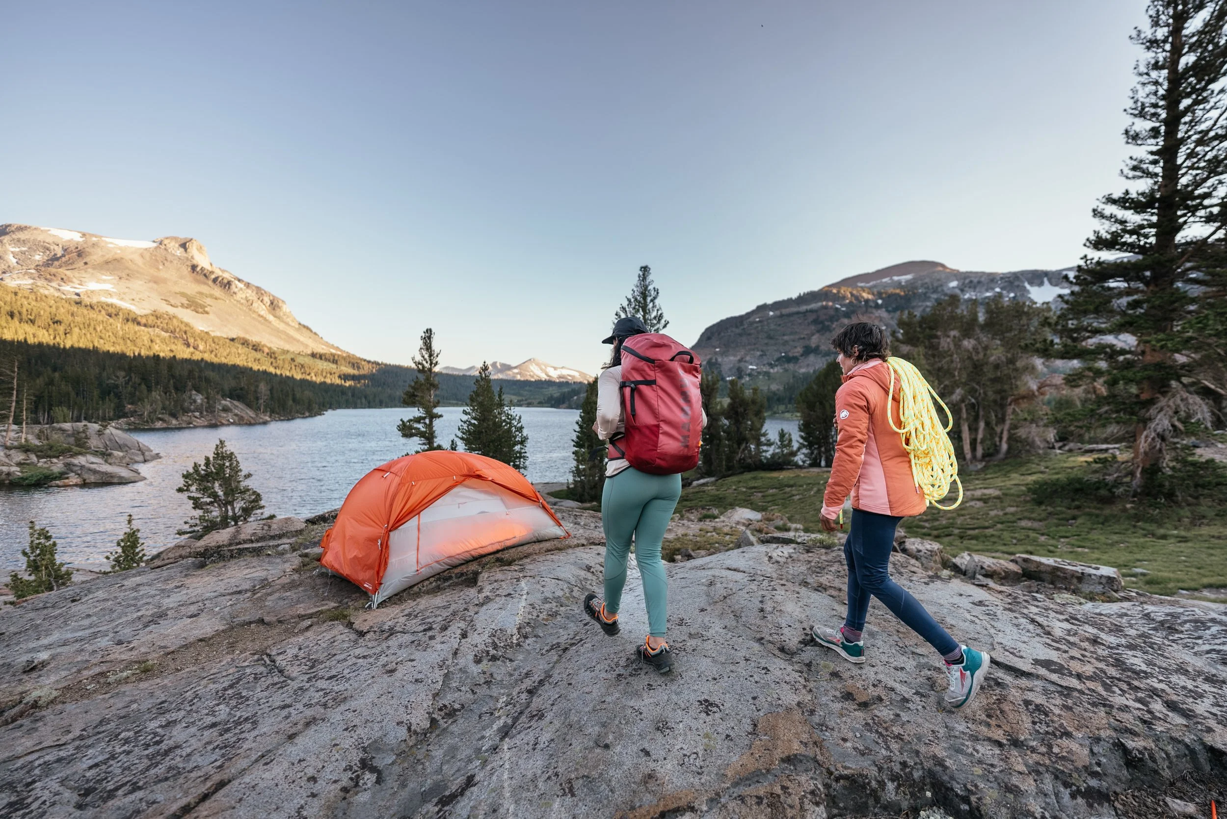 Two women hiking in a mountainous area near a lake, with a small orange tent set up on a rocky surface, surrounded by trees and mountains in the background.