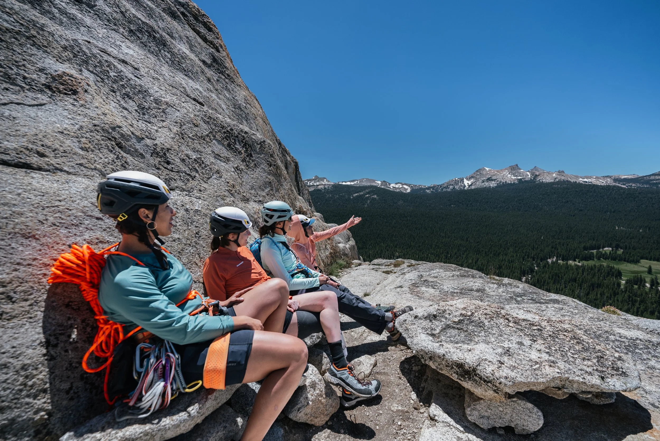 Group of women in helmet and outdoor gear resting on a rock ledge with a mountain view.