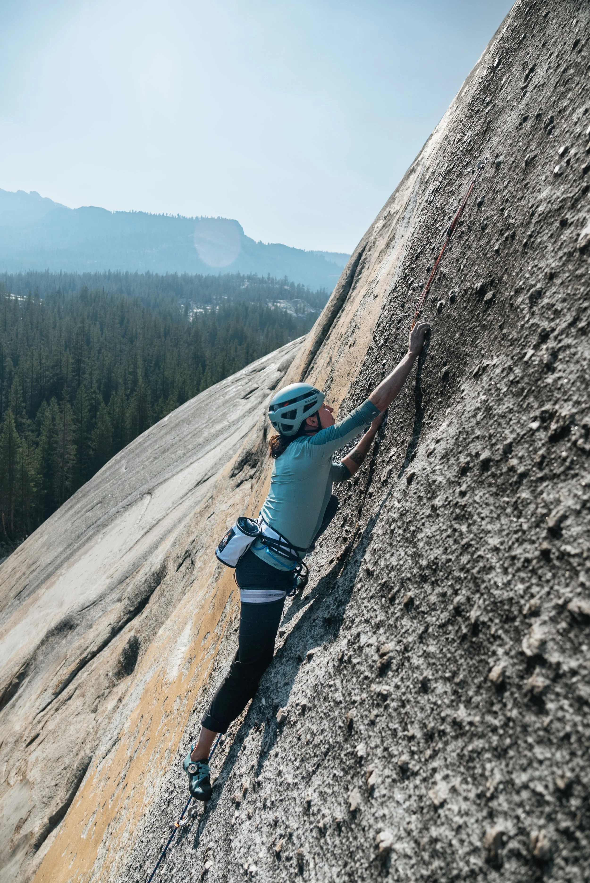 A woman rock climbing on a steep granite cliff, wearing a helmet and climbing gear, with a scenic forest and mountain landscape in the background.