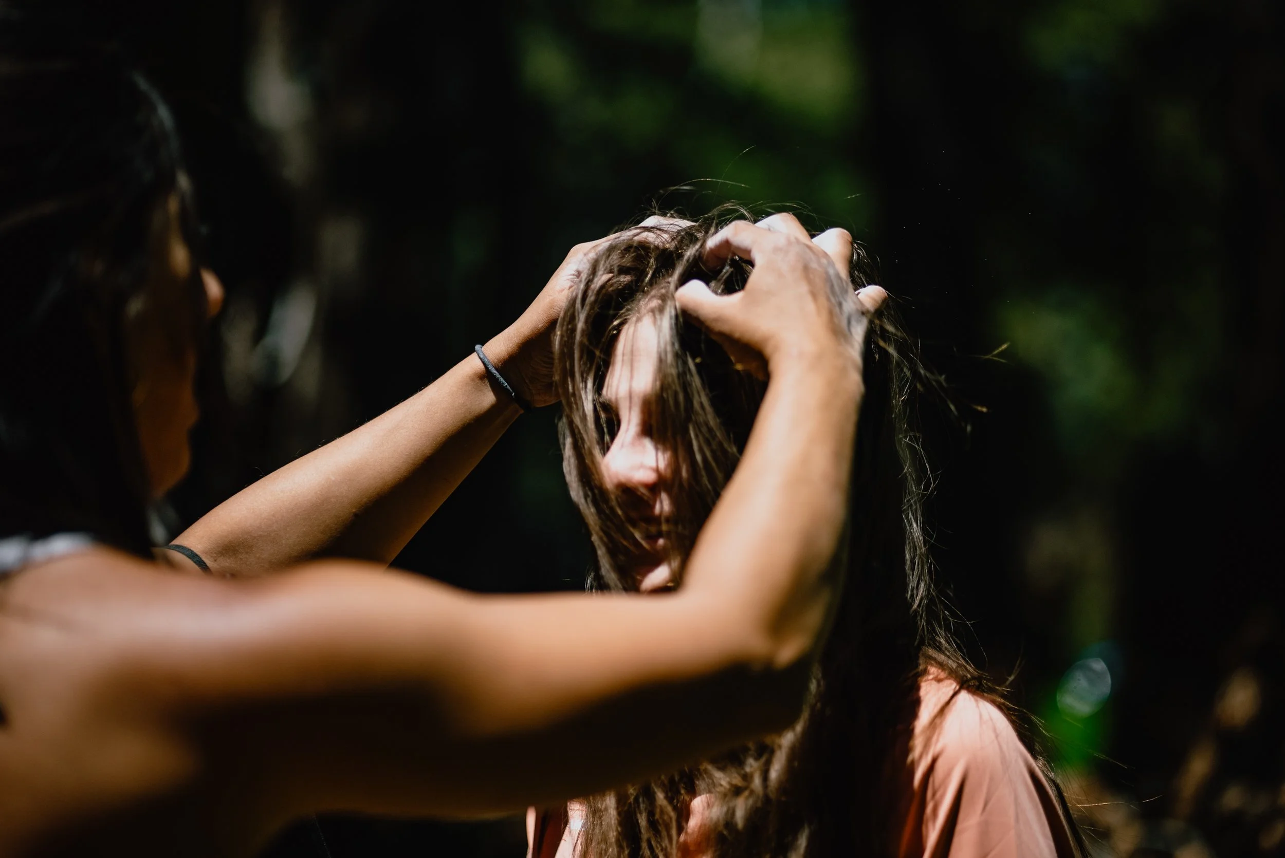 Two women are outdoors, one touching the other's hair in a gentle, caring manner. The scene appears emotional or intimate.