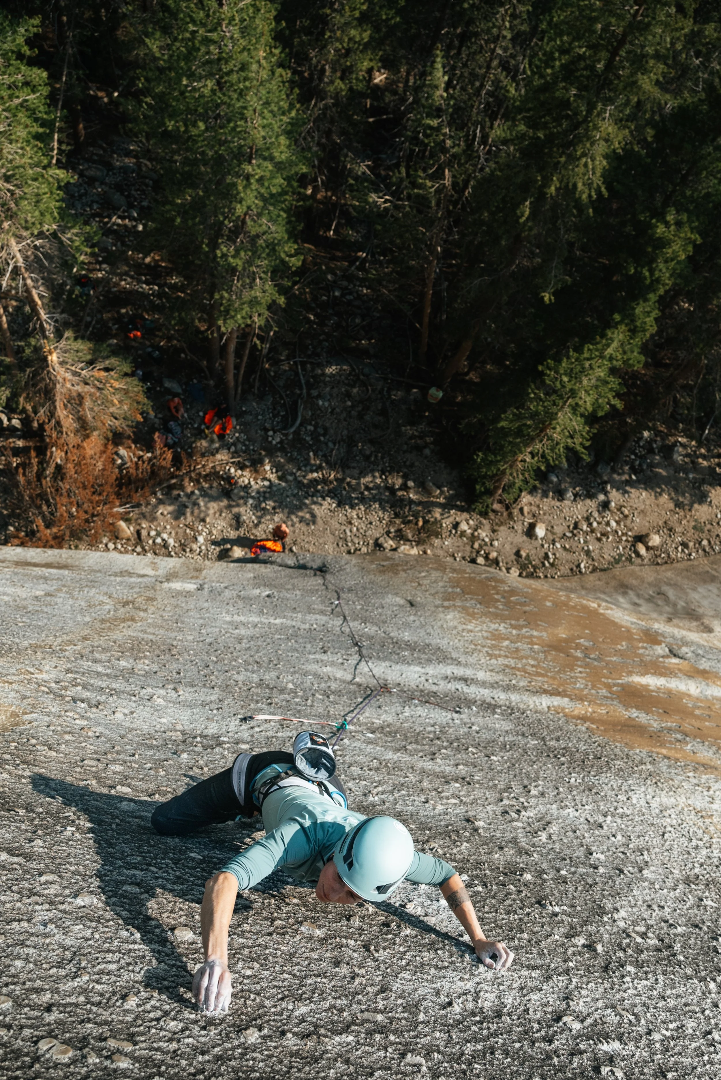 A person rock climbing on a steep outdoor rock face, wearing a helmet, harness, and climbing gear, with trees visible at the base.