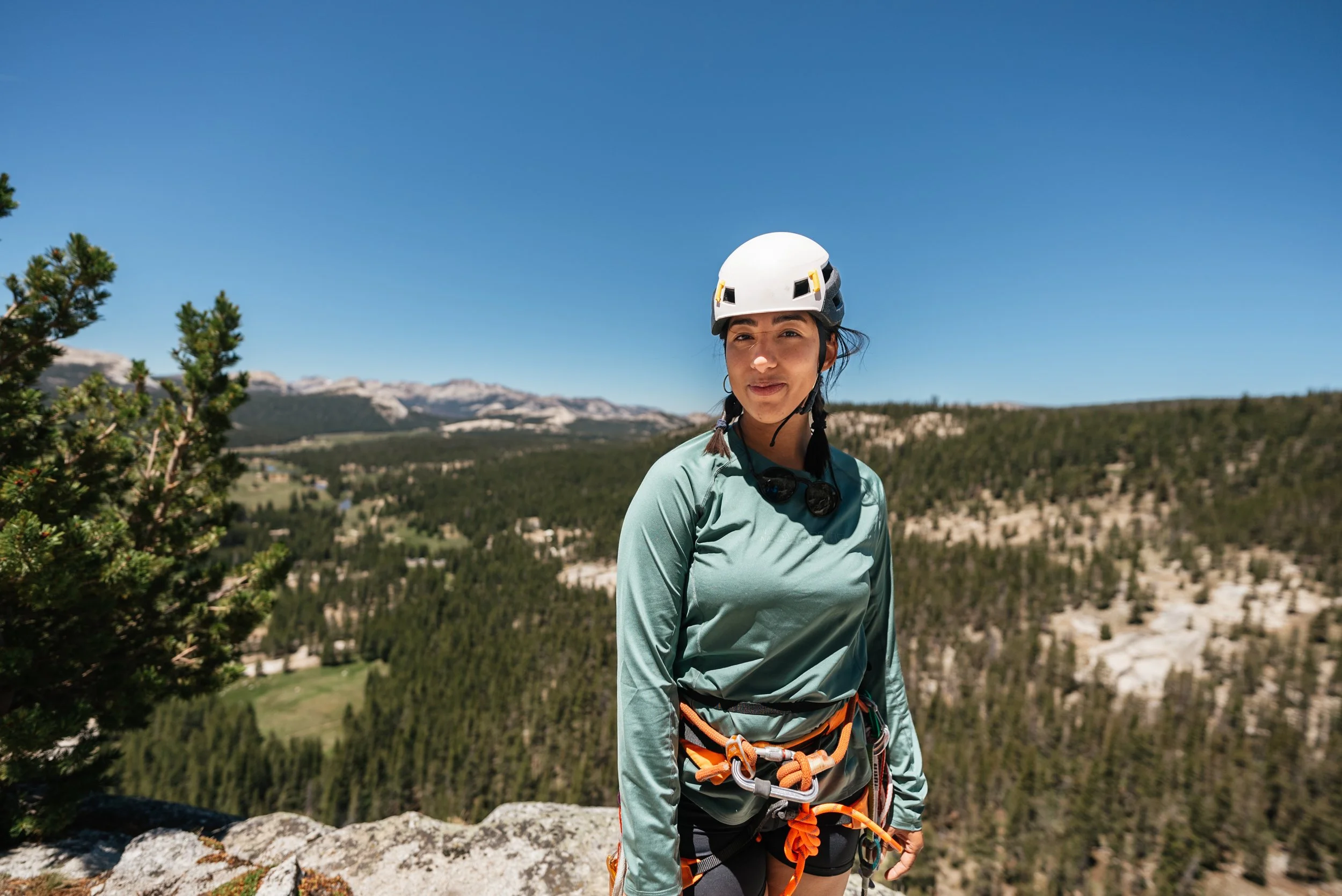 Woman in outdoor gear with helmet and harness standing on rocky edge overlooking forested mountains under clear blue sky.