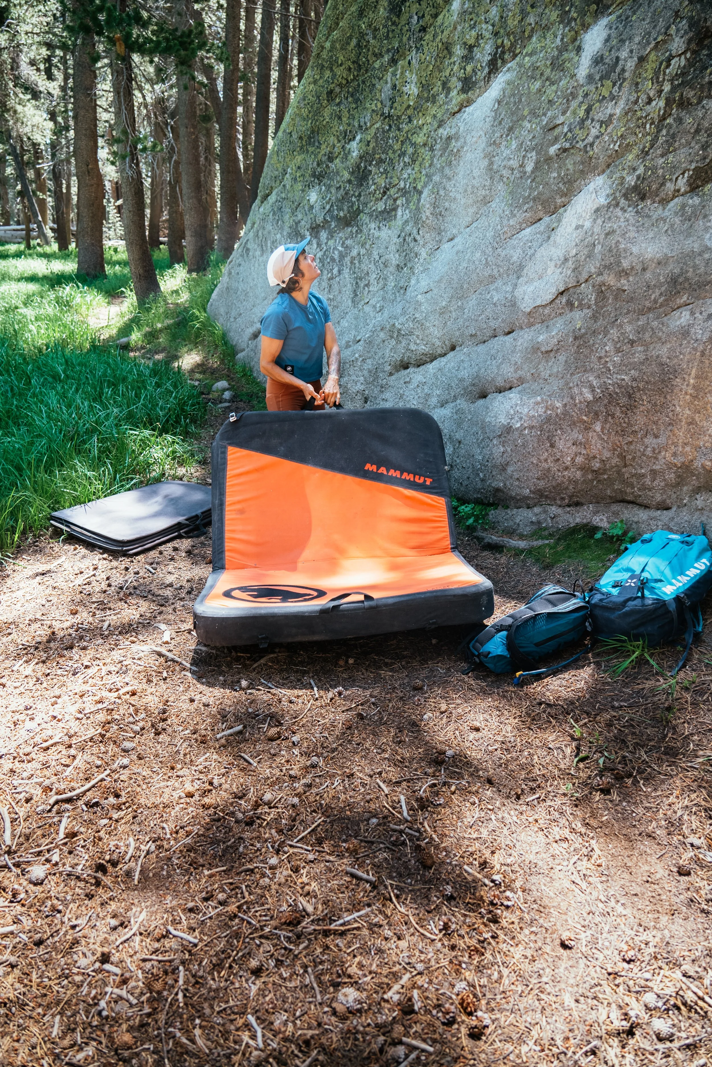 A woman in outdoor gear standing near a large rock formation in a forest, with climbing gear and pads nearby.
