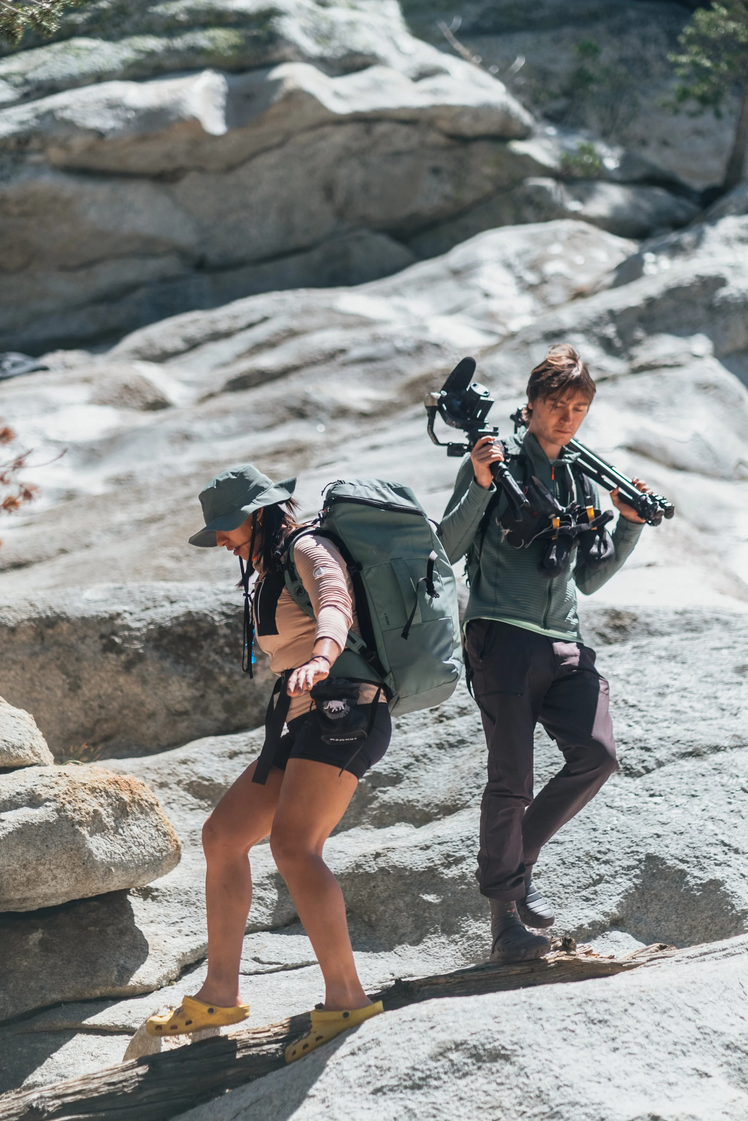Two hikers crossing a rocky terrain, with one carrying a large backpack and the other holding a camera and tripod.