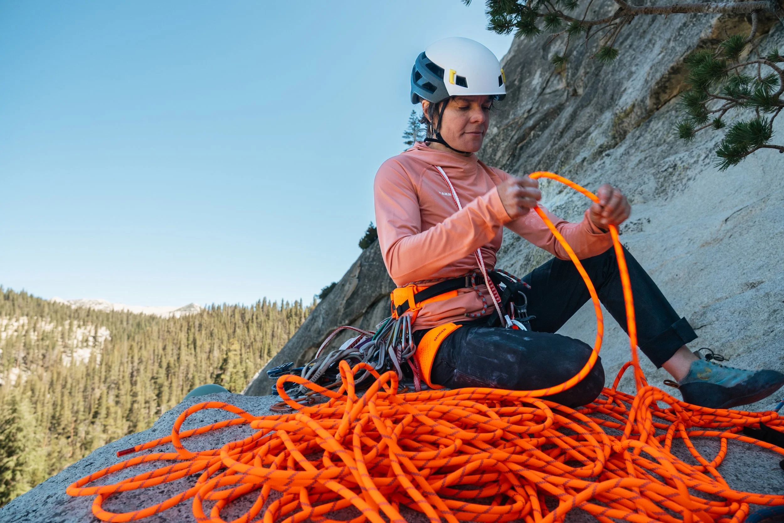 A woman wearing a helmet and climbing gear sitting on a rock while preparing climbing ropes outdoors.