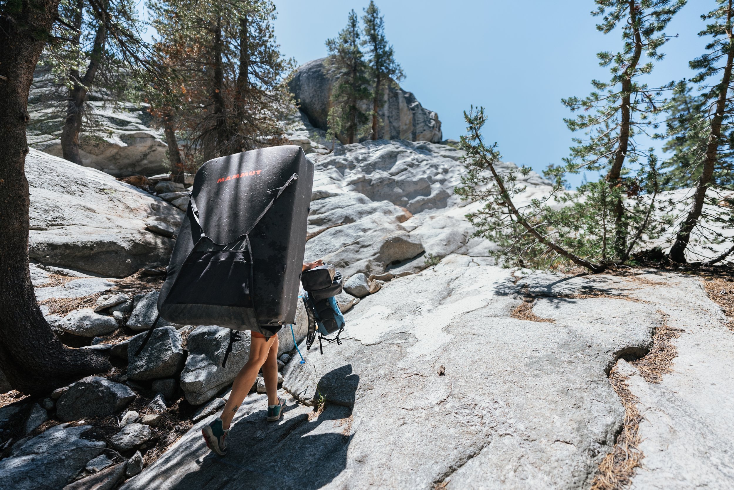 A person climbing a rocky mountain trail carrying a large black Mammut climbing or camping gear bag on their back, with trees and a clear blue sky in the background.