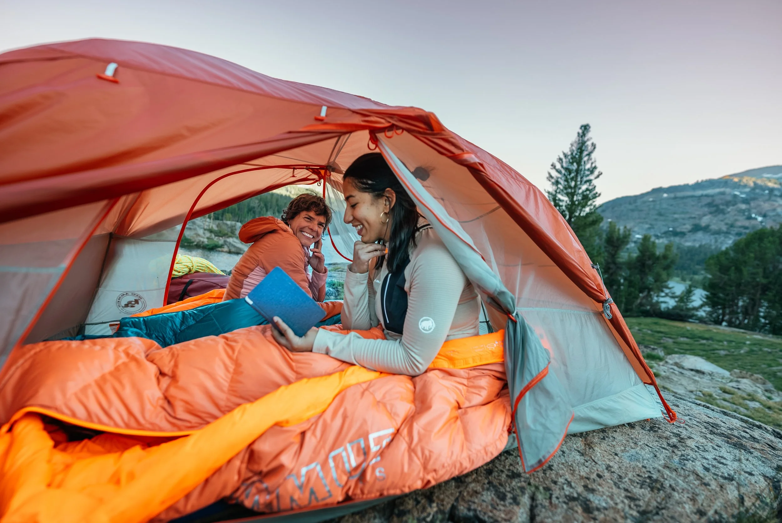 Two women inside an orange camping tent outdoors, laughing and reading a book, with trees and mountains in the background during daytime.