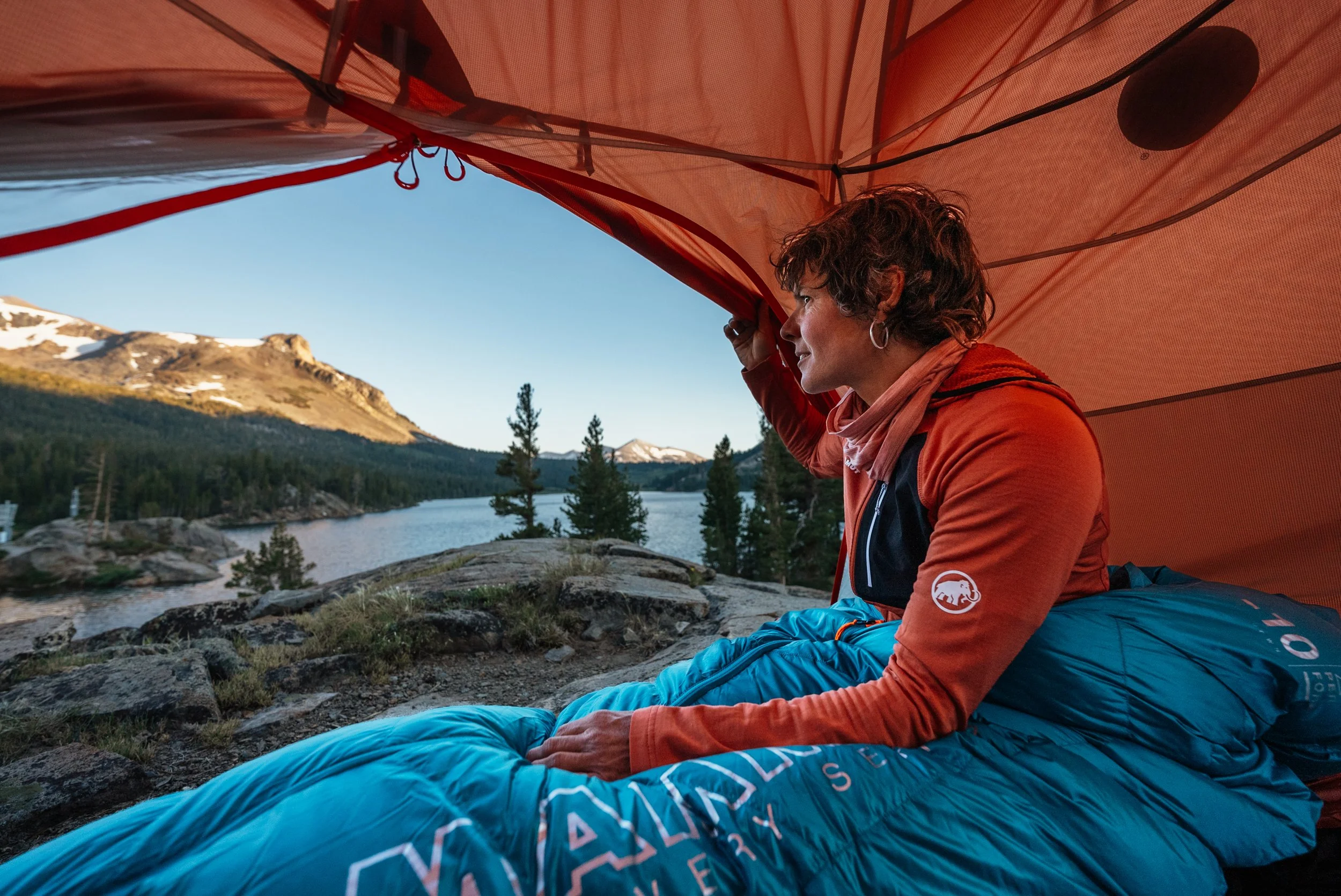 A person inside a red tent, sitting on a blue sleeping bag, overlooking a scenic mountain lake with pine trees and snow-capped peaks in the background.