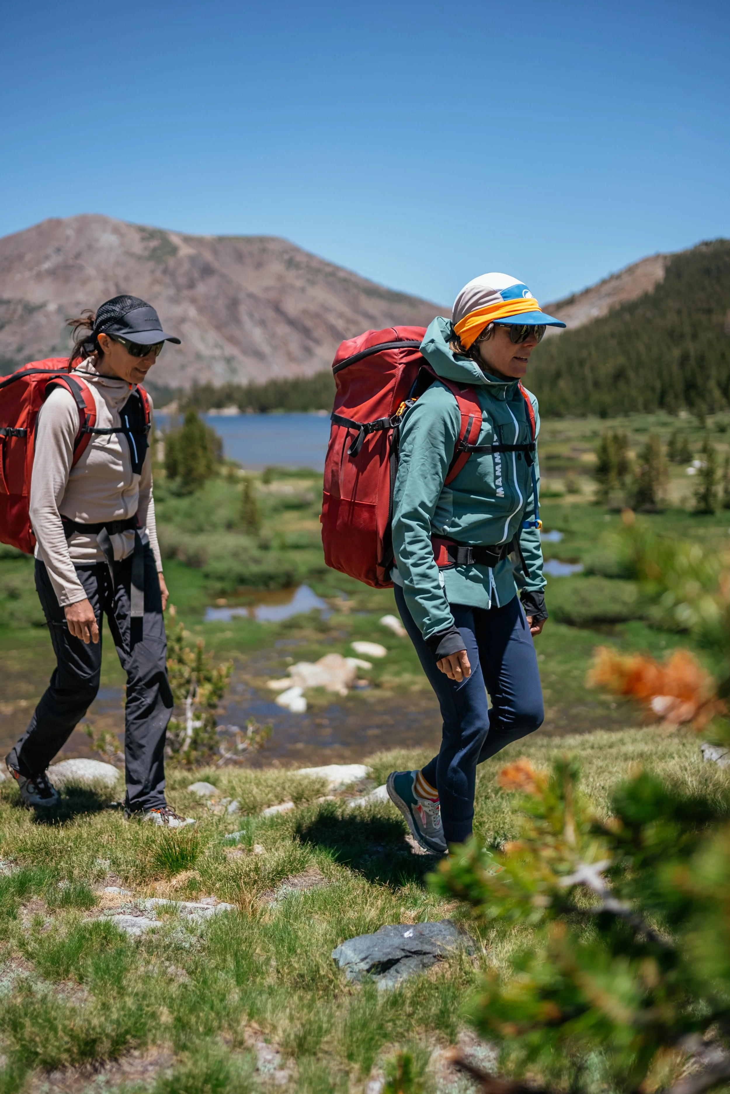 Two women hiking with backpacks in a mountainous landscape with a lake and green trees.