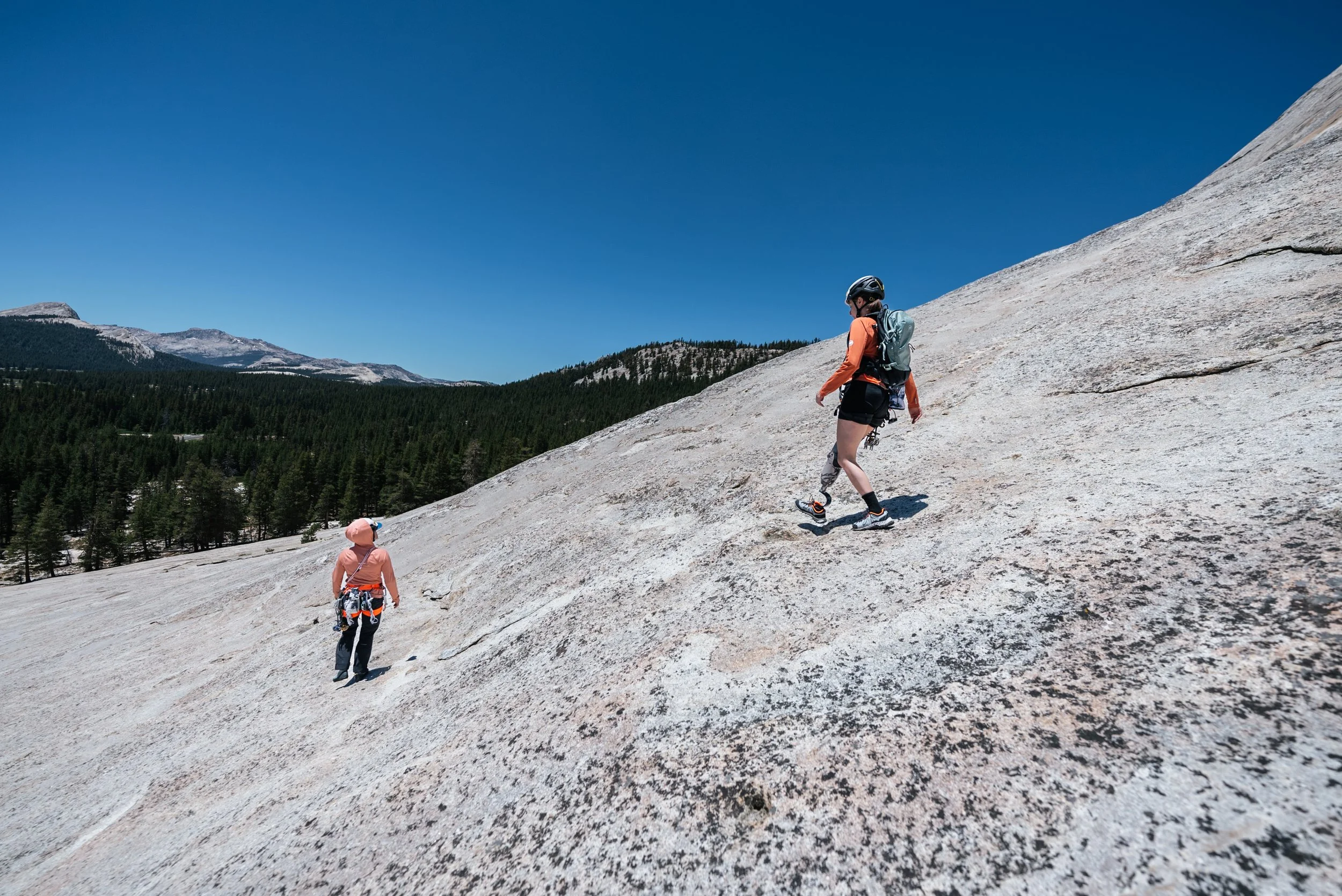 Two hikers climbing a large, sloped granite rock face in a mountainous landscape, with pine trees and distant mountains under a clear blue sky.