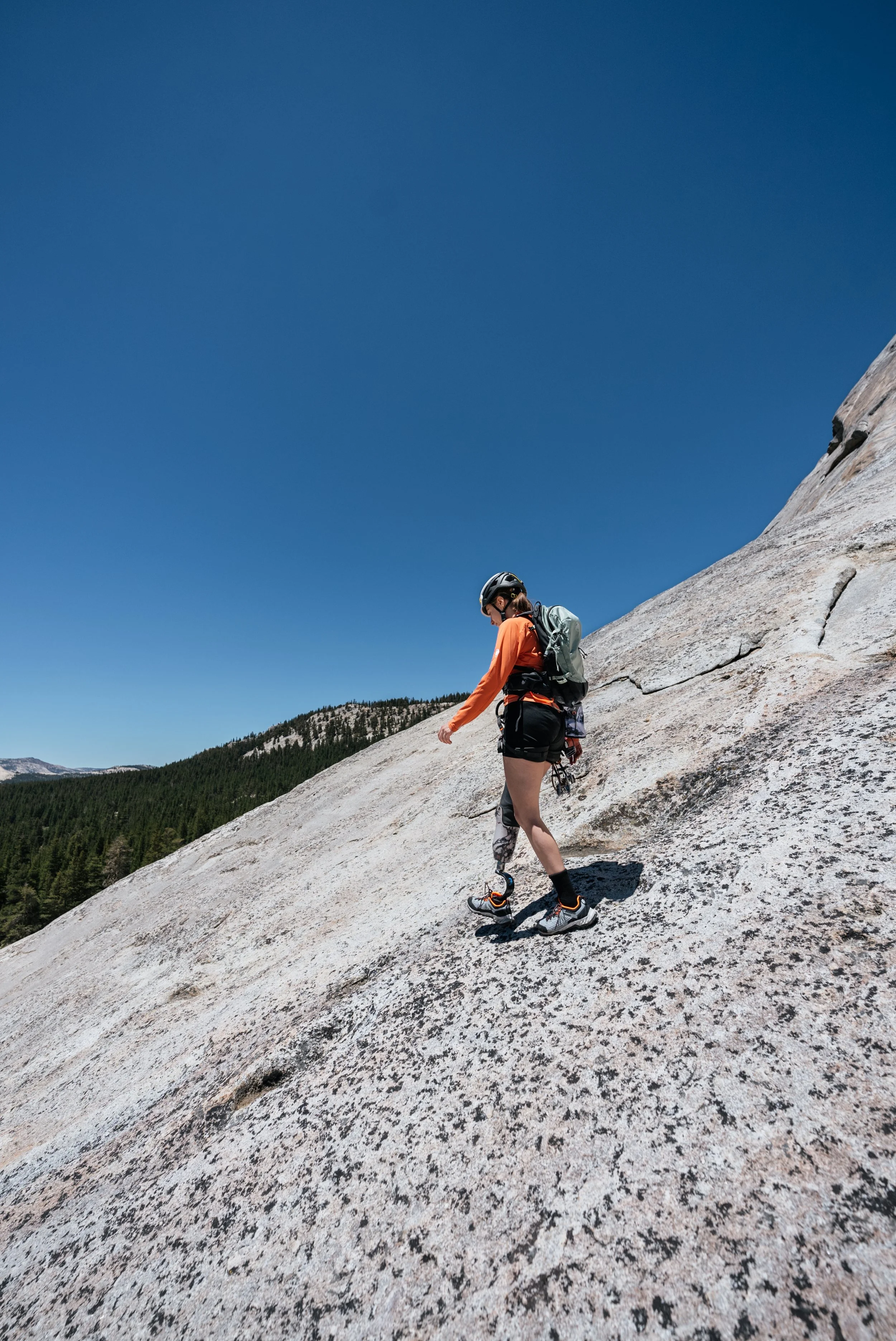 A woman climbing a steep granite slope in a mountainous area, wearing climbing gear, helmet, and backpack under a clear blue sky.