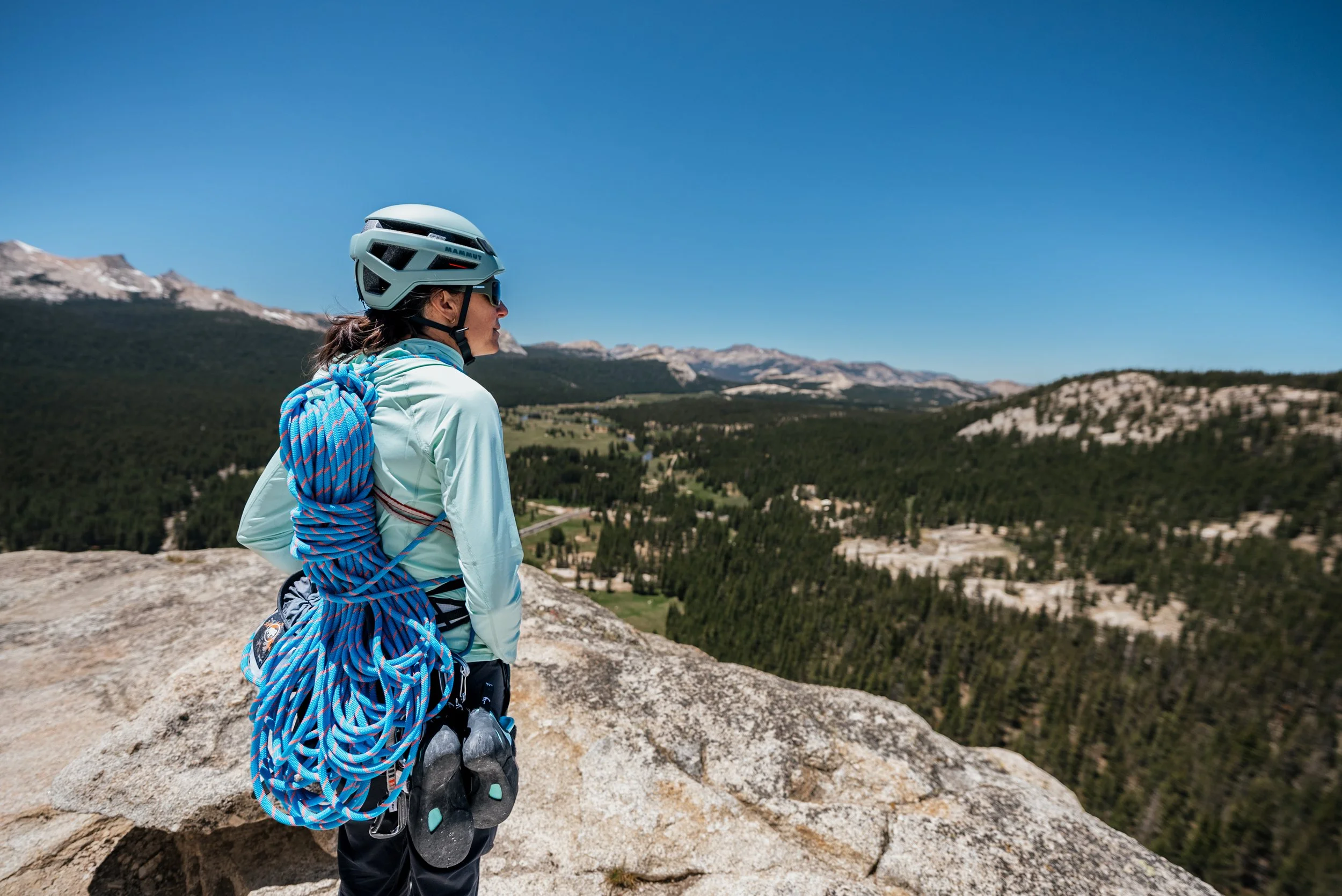 Female climber with a helmet and sunglasses standing on a rock overlooking a forested landscape with mountains in the distance during daytime.