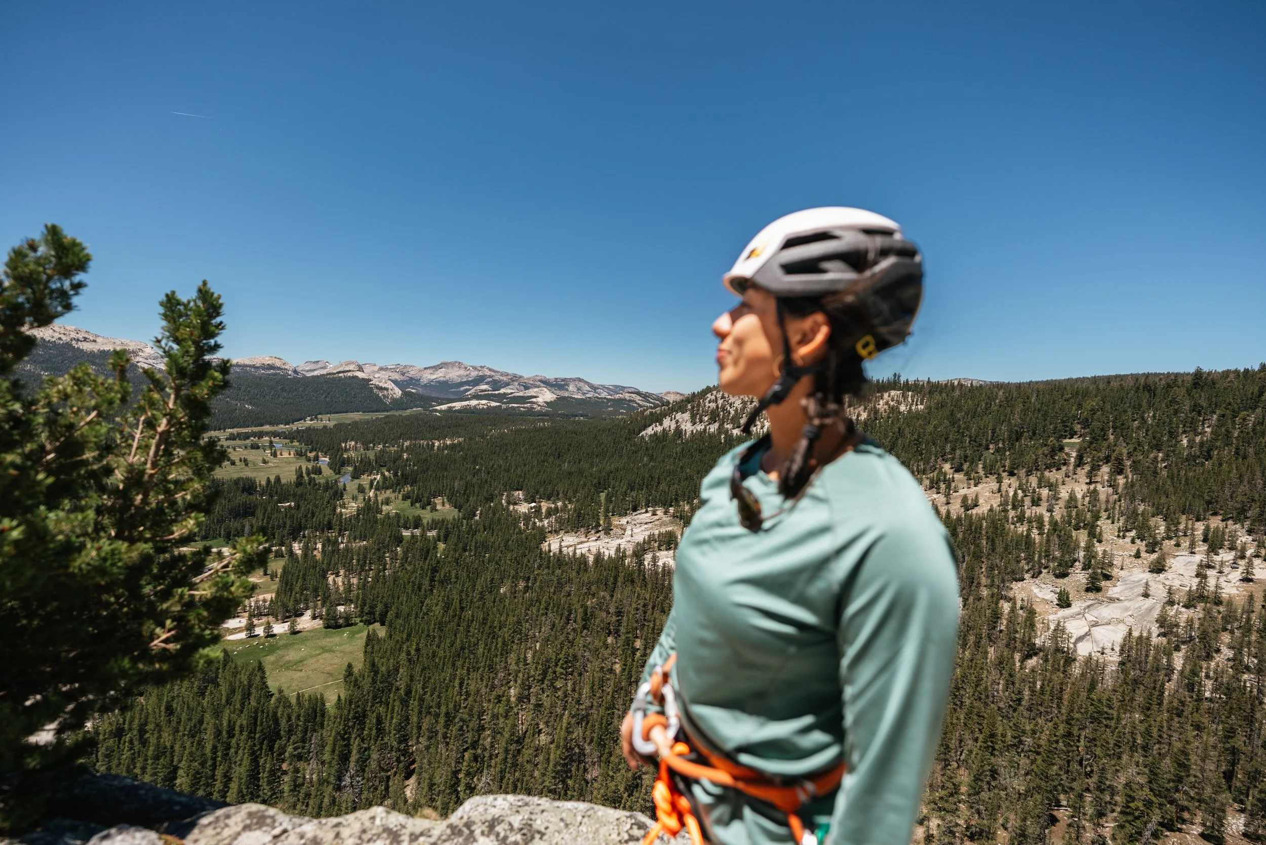 A woman in a helmet and outdoor gear stands on a rocky outcrop overlooking a forested mountain landscape under a clear blue sky.